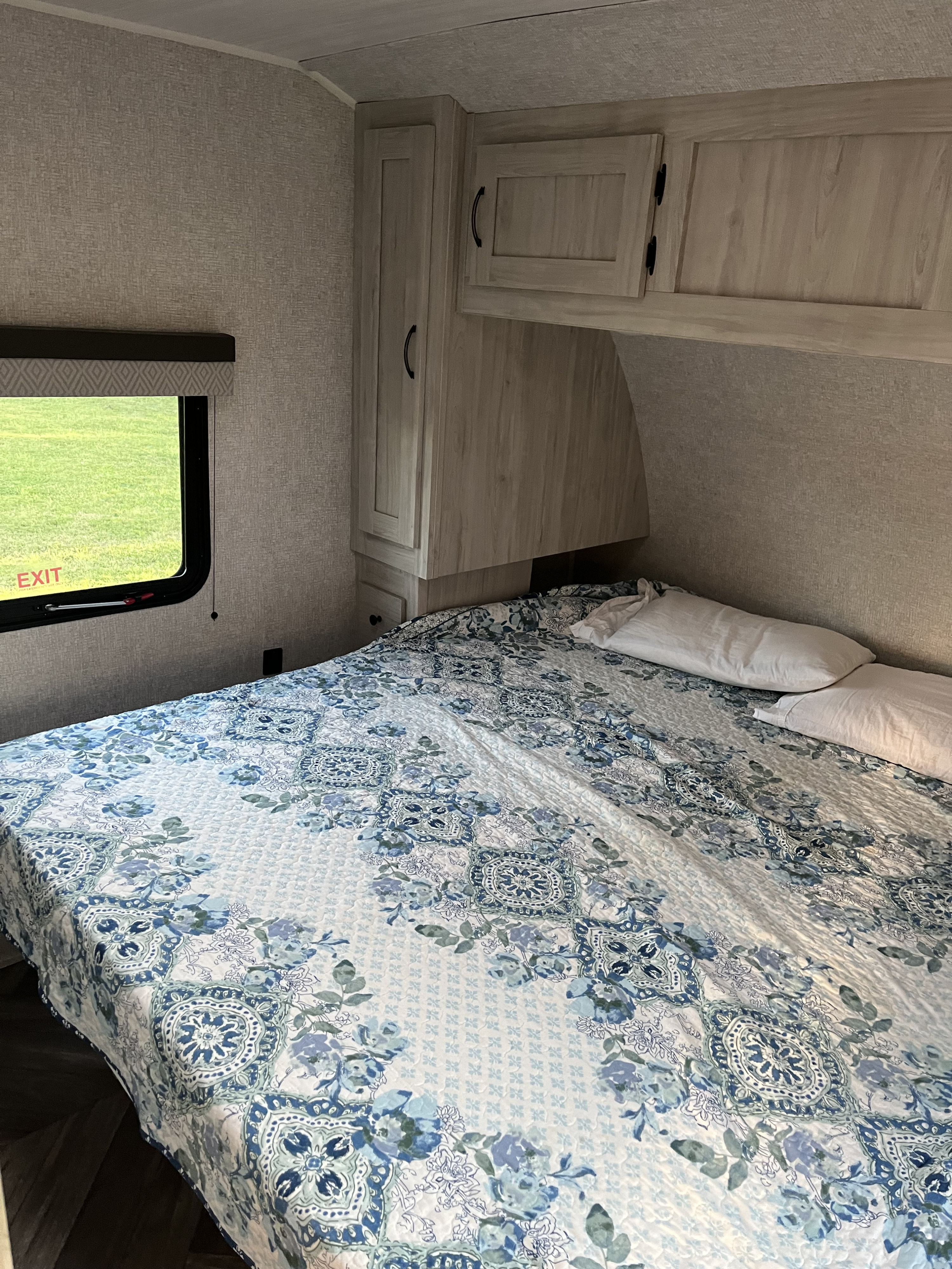 Cozy RV rental bedroom with floral bedding, wall cabinets, and a window overlooking a green field, perfect for travel.. East to West Della Terra 2022