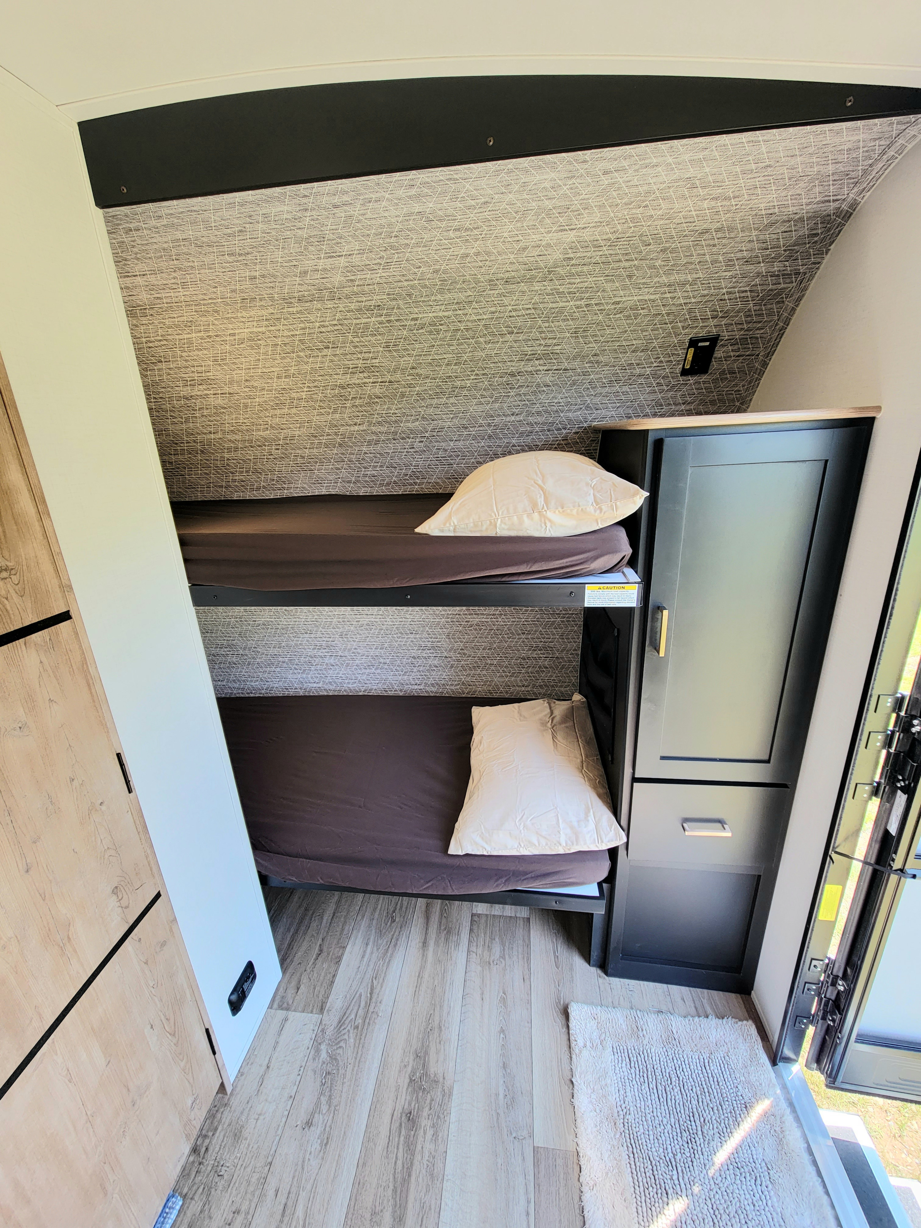 Interior view of an RV rental showing compact bunk beds with pillows & dark sheets, alongside a small cabinet.. Keystone Outback 2023