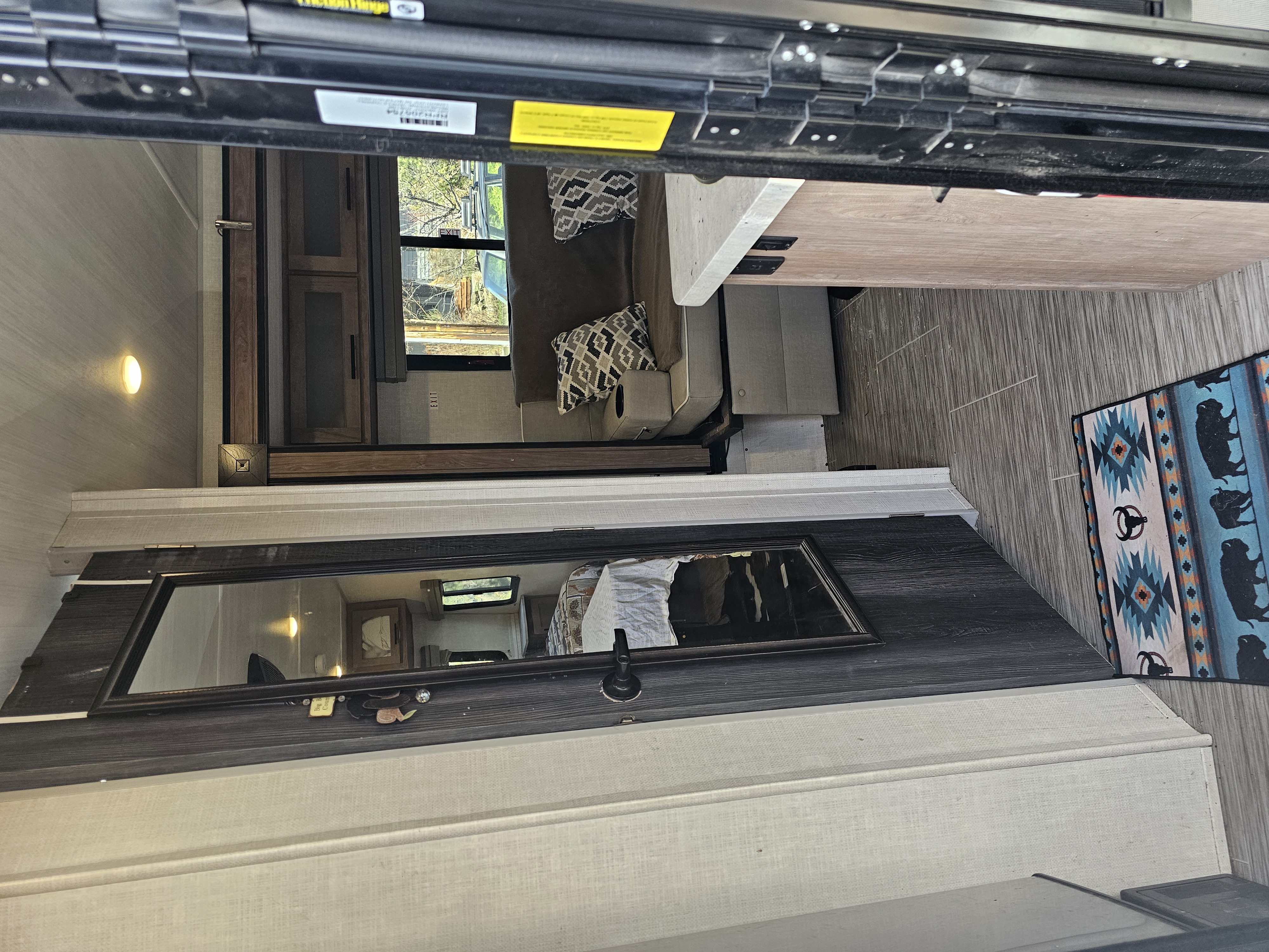 Inside a modern RV rental: a bright living space with a sofa, wood-look floor, and large window offering nature views. Reflective surfaces add depth.. No Boundaries 19 2021