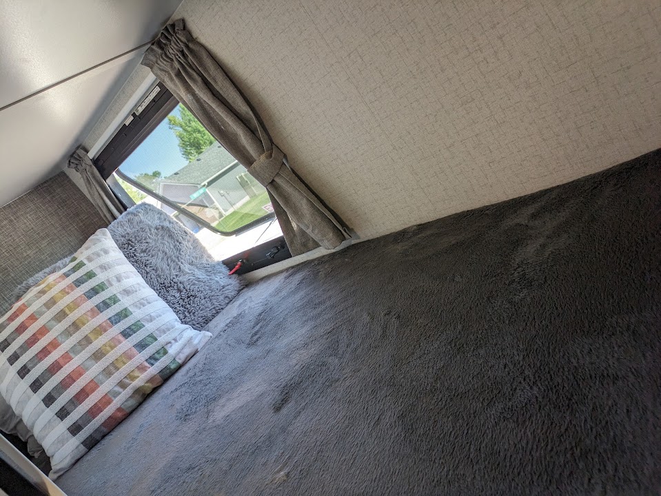 Inside an RV rental bunk: plush grey bedding, striped & fluffy pillows, with a window showing houses. Cozy travel space.. Winnebago Micro Minnie 2022