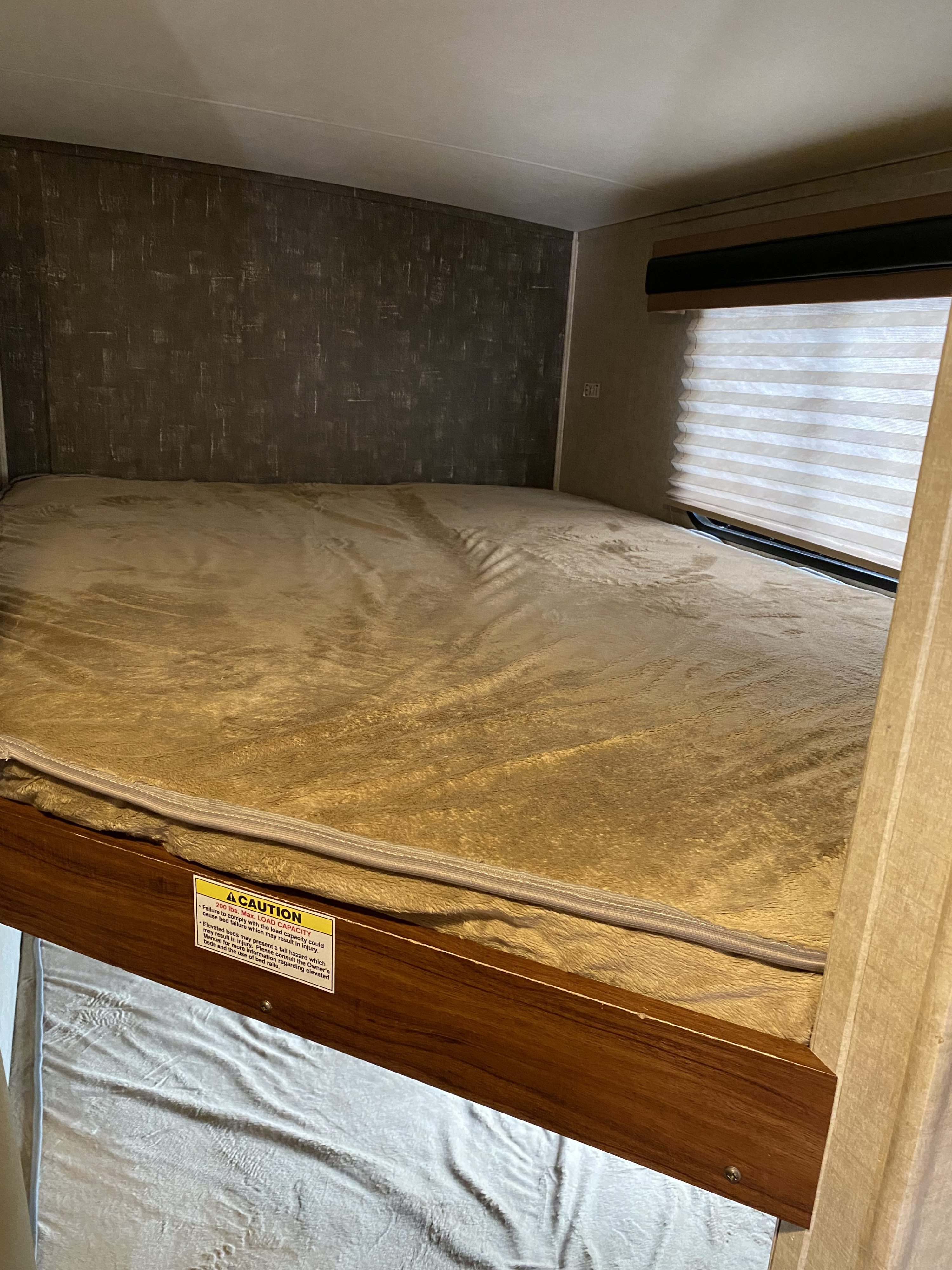 Elevated bed in an **rv rental** showing a 200 lbs. max load safety sign and cozy blanket, with window blinds.. Heartland RVs North Trail 2016