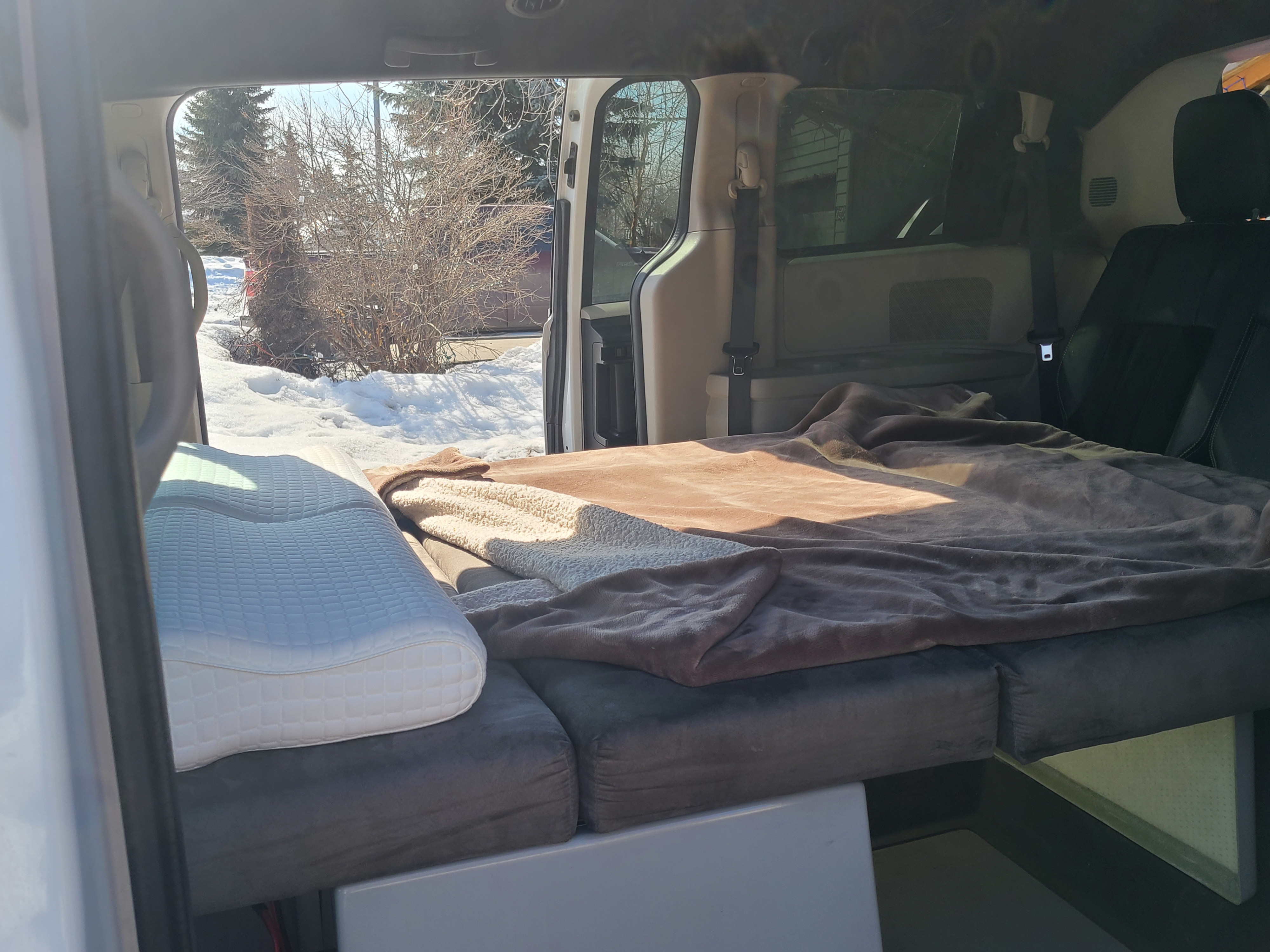 Cozy van conversion setup for an RV rental, featuring a bed, pillow, and blanket, with a snowy outdoor view.. Dodge Mini C 2017