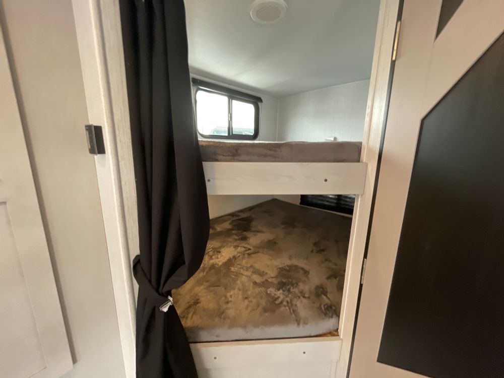 This RV rental features compact bunk beds: a cozy sleeping area with two mattresses, a window, and a tied-back curtain, ideal for travel.. Heartland RVs Mallard 2023
