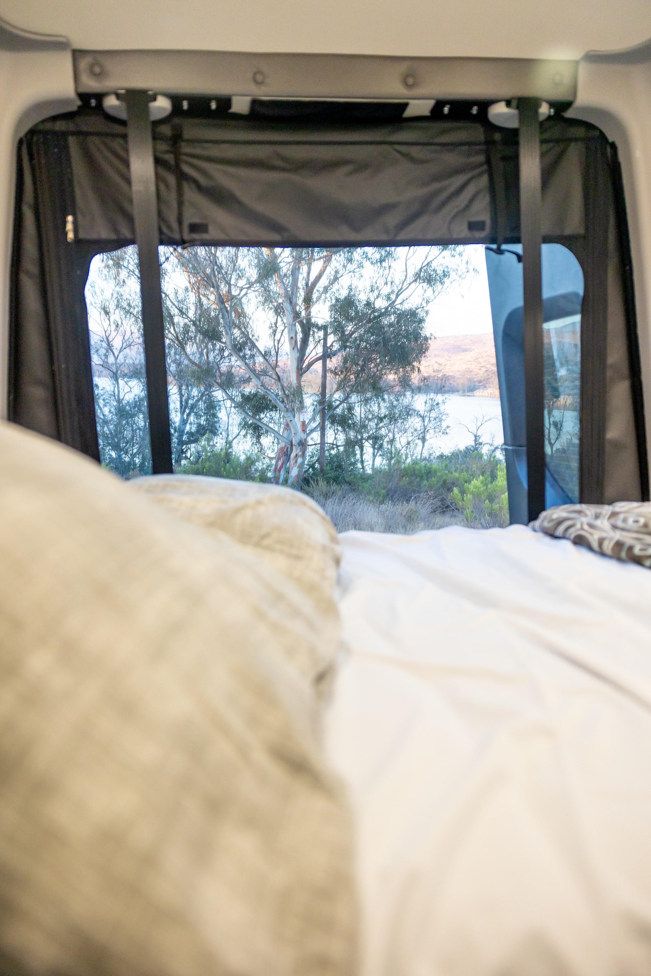 Cozy RV rental interior: a blurred bed leads to a clear view of a tranquil lake, trees, and distant hills.. Winnebago Revel 2023