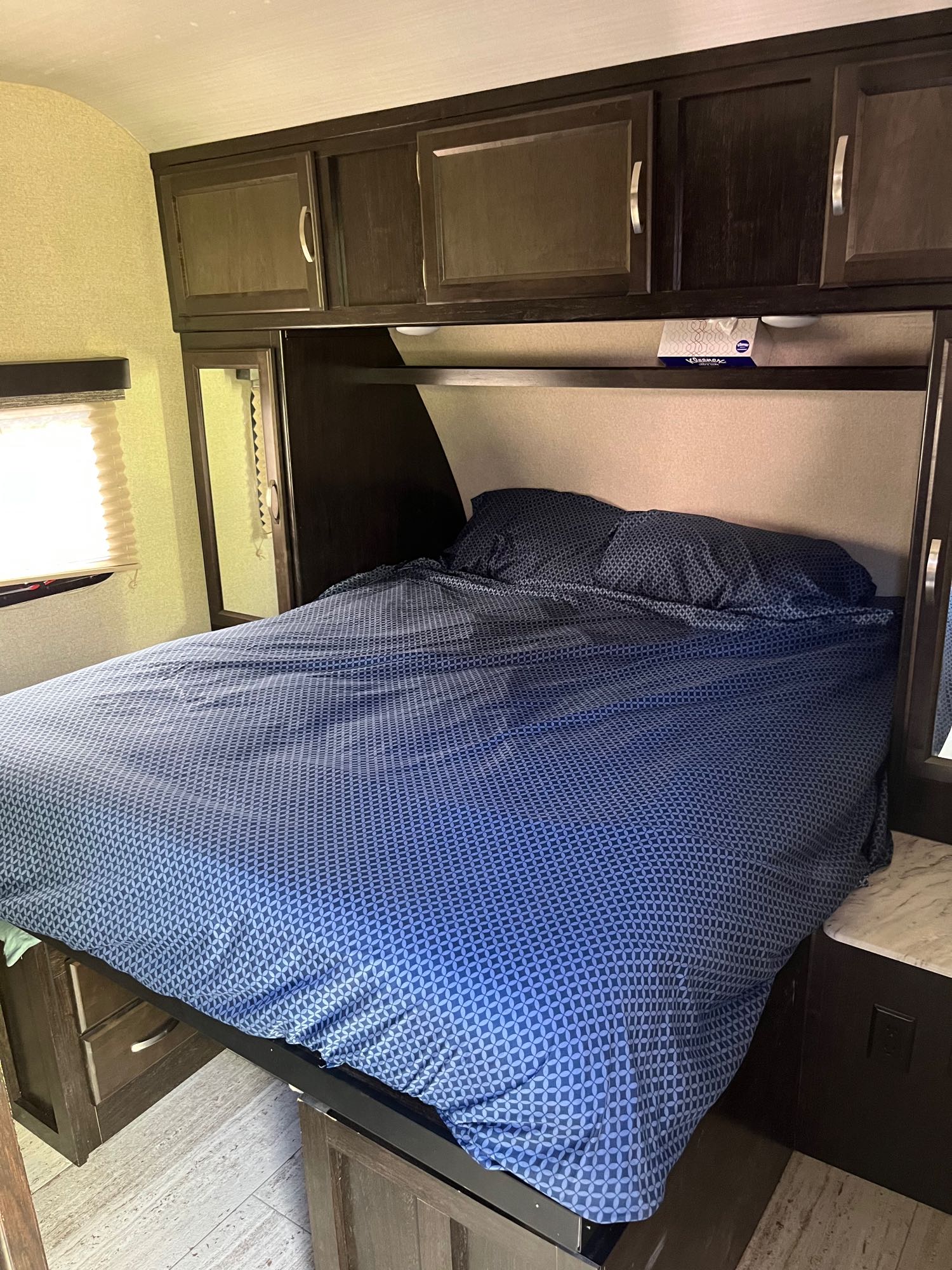 Cozy RV rental bedroom featuring a bed with blue geometric bedding, overhead cabinets, and a window.. Coachmen 2963bh 2020