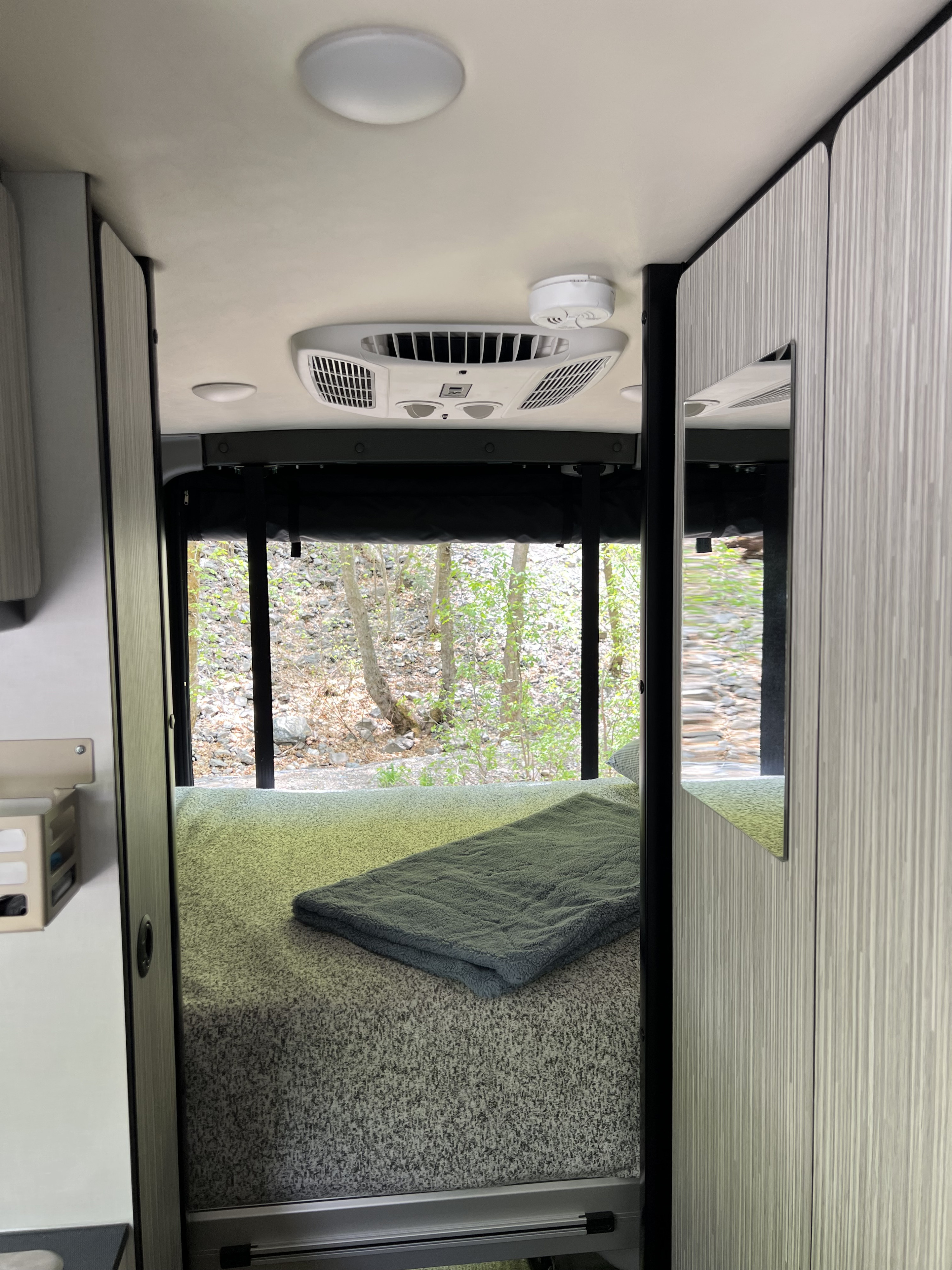 Inside an RV rental: a cozy bedroom with large window overlooking nature, featuring AC, lighting, and storage.. Winnebago Revel 2023