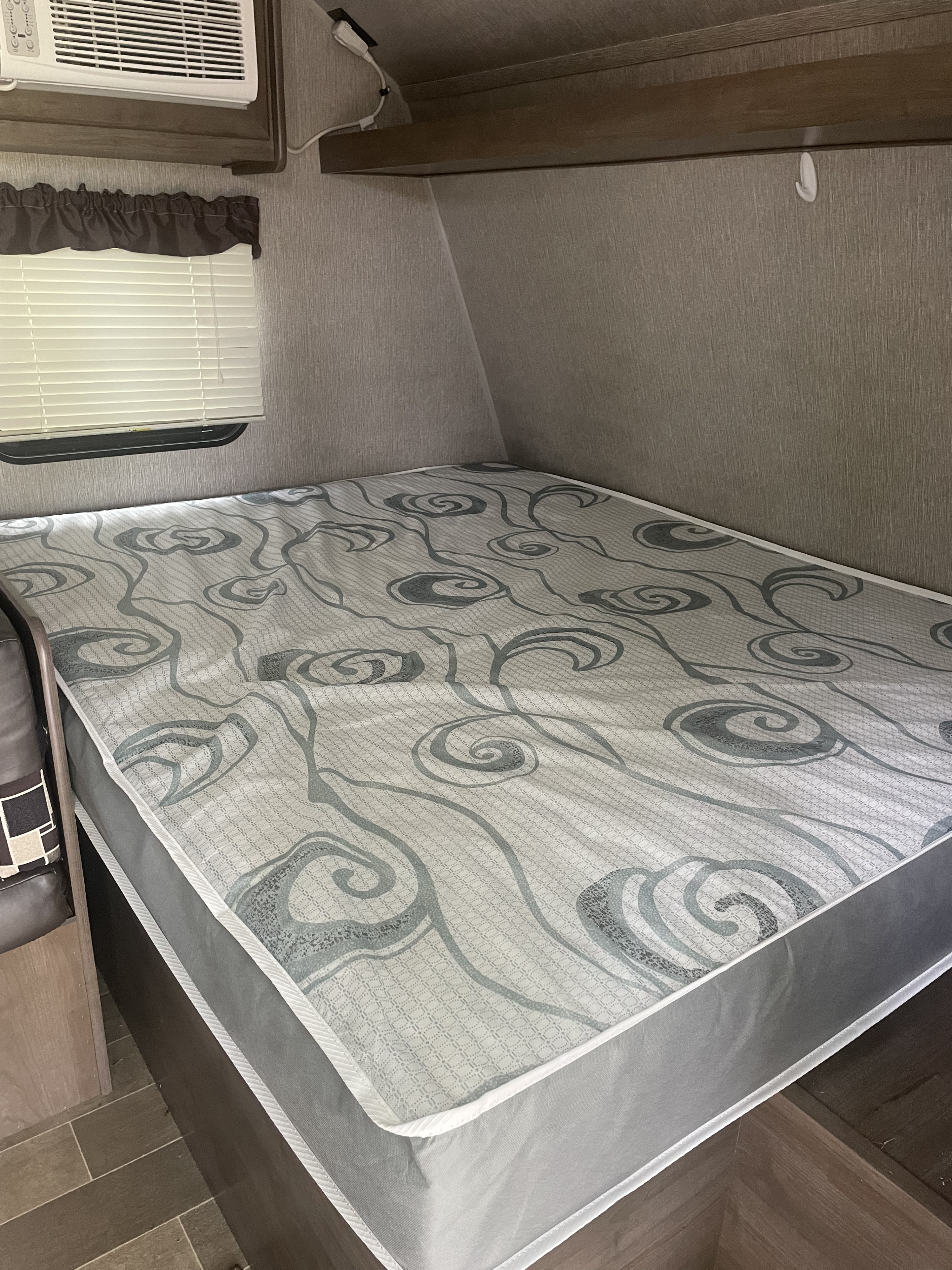 Cozy RV rental interior: view of a bed with a gray patterned mattress, window with blinds, and mounted AC unit.. Dutchmen Coleman Lantern LT 2019