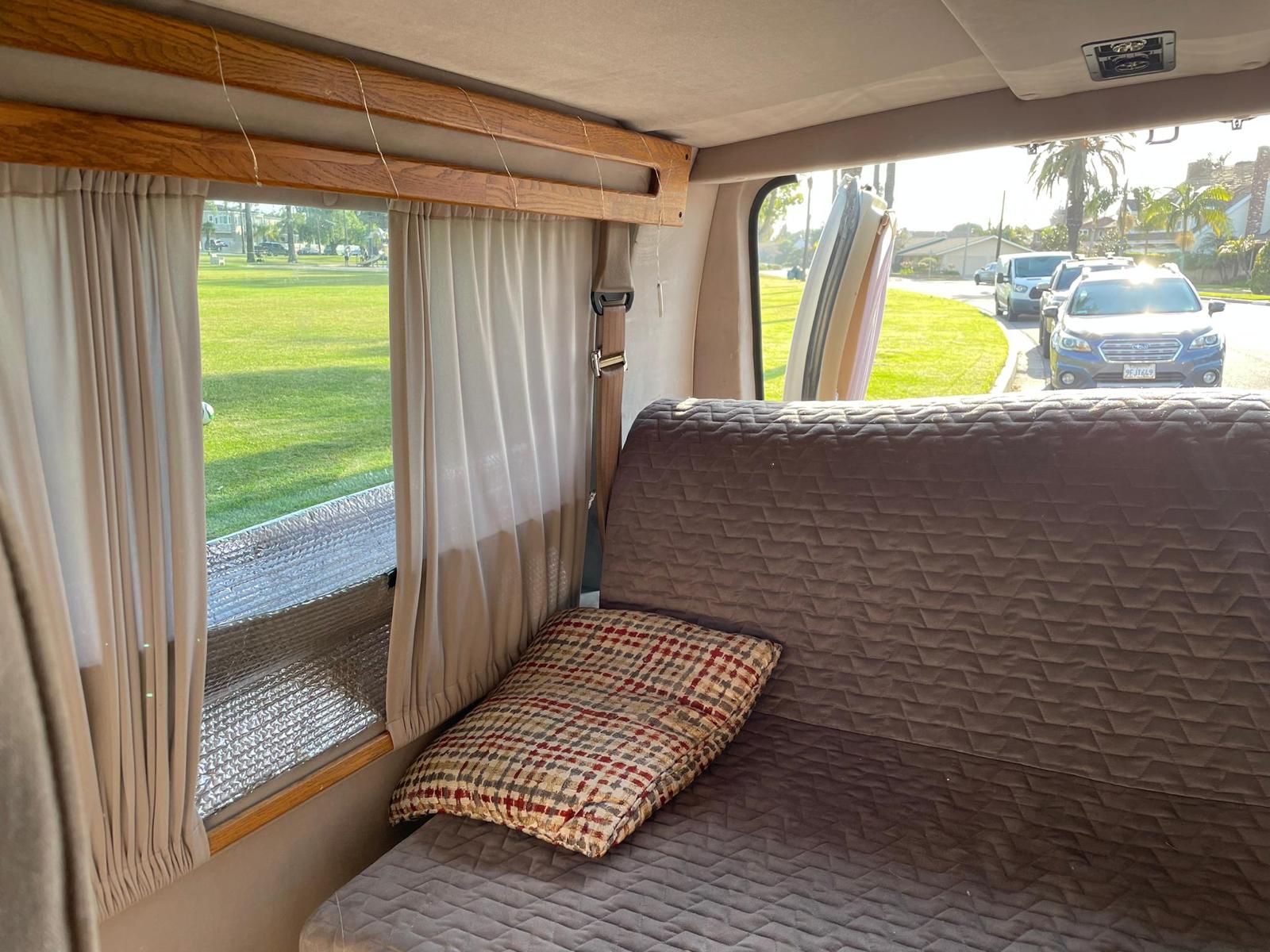 RV rental interior featuring a comfy quilted bed, plaid pillow, window curtains, and a sunny park view.. Ford SV 1994