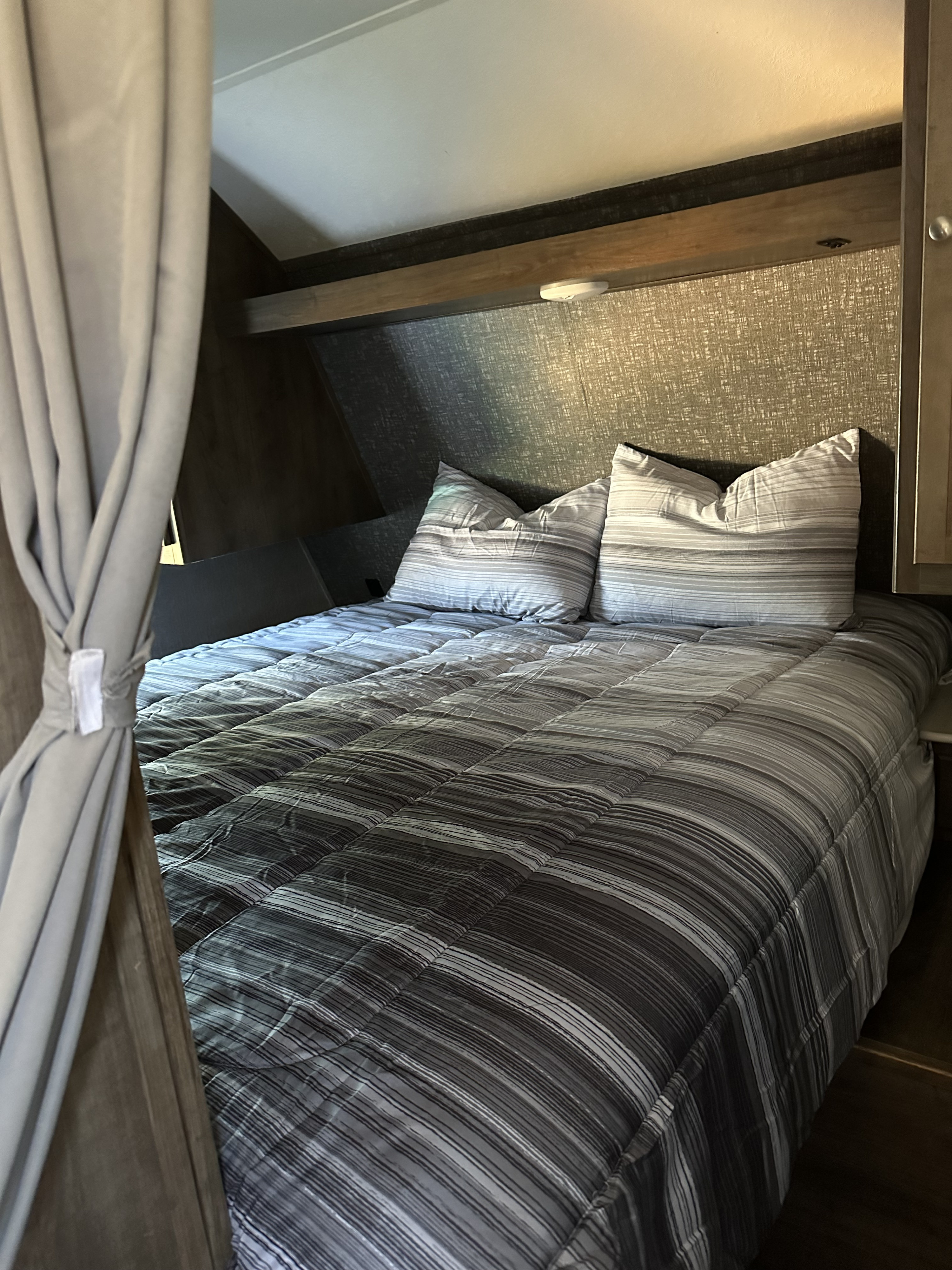 Cozy RV rental bedroom: A comfortable bed with gray and white striped bedding, pillows, and a light curtain tied back.. Dutchmen Coleman Lantern 2020