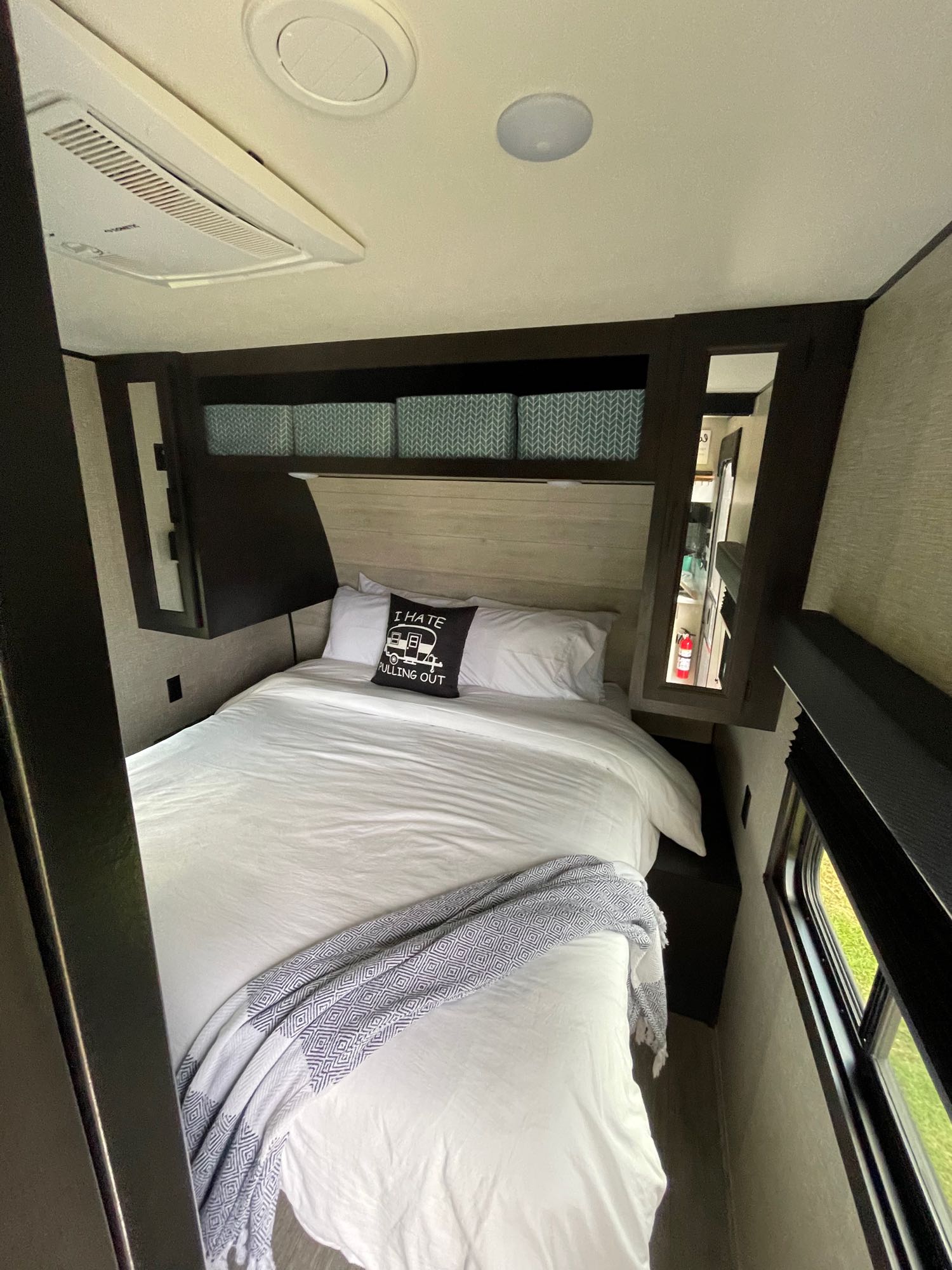 Comfortable RV rental bedroom: queen bed with white bedding, AC unit, overhead storage, and window showing grass. Features a humorous camper pillow.. Jayco Jayflight 324BDS 2023