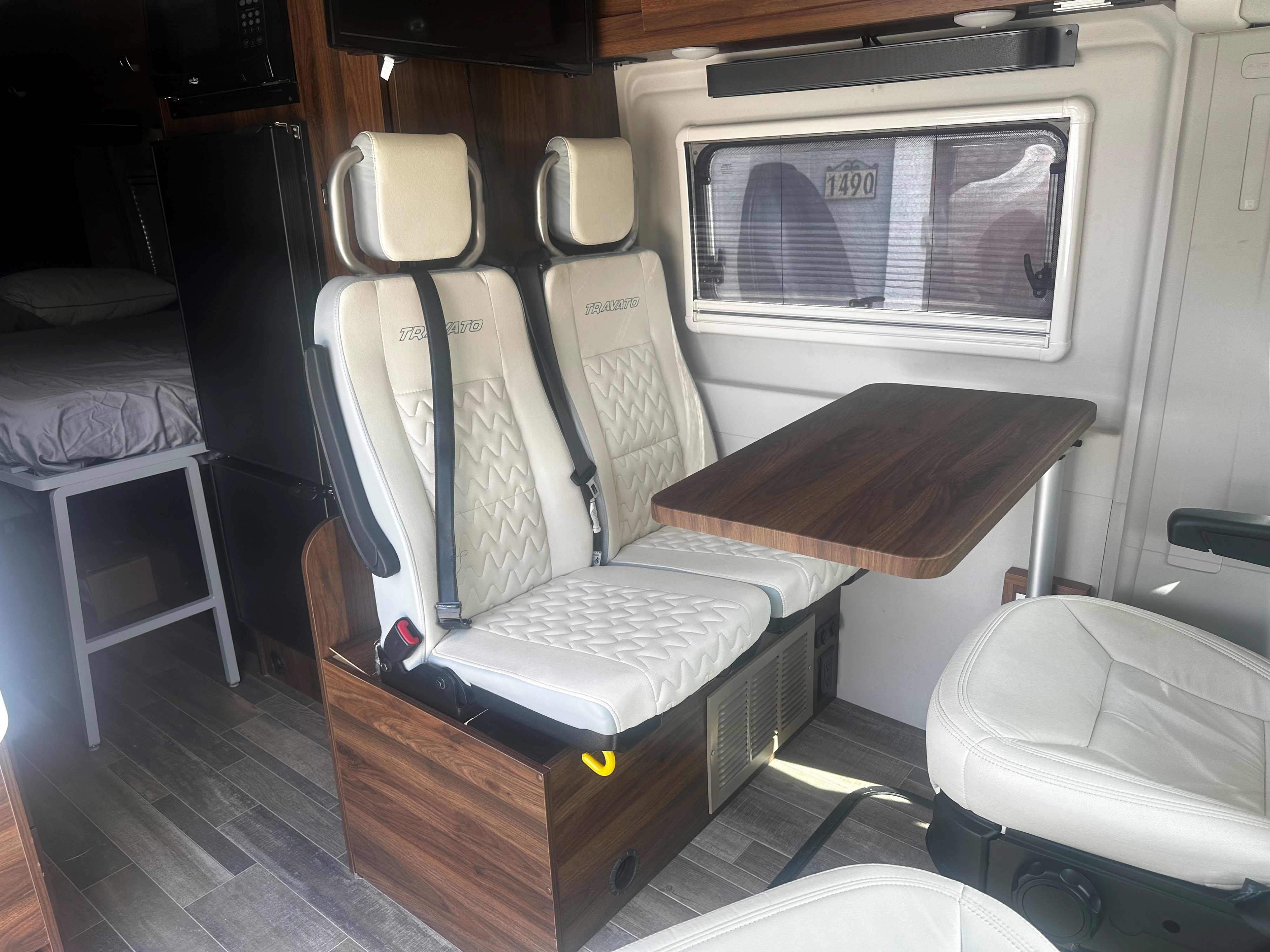 This RV rental interior features white leatherette seats, a dining table, and a cozy rear bed, perfect for travel adventures.. Winnebago Travato 2022