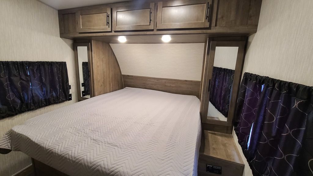 RV rental bedroom with a large bed, light quilted cover, overhead wood cabinets, and dark patterned curtains. Features side mirrors.. Forest River Cherokee Wolf Pack 2021
