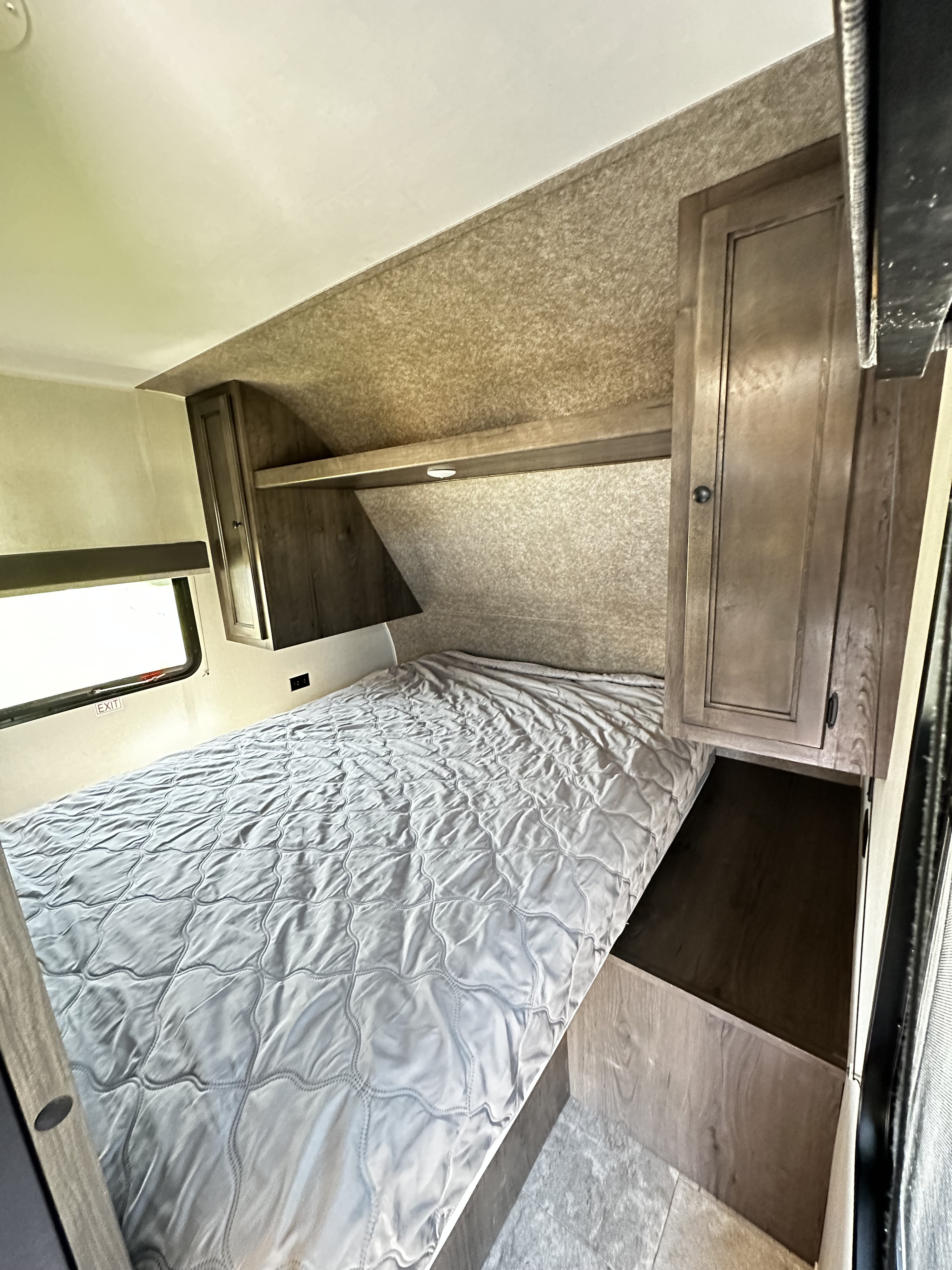 Cozy RV rental bedroom featuring a gray quilted bed, overhead shelf, window, and wood-look storage.. Starcraft Autumn Ridge 2021