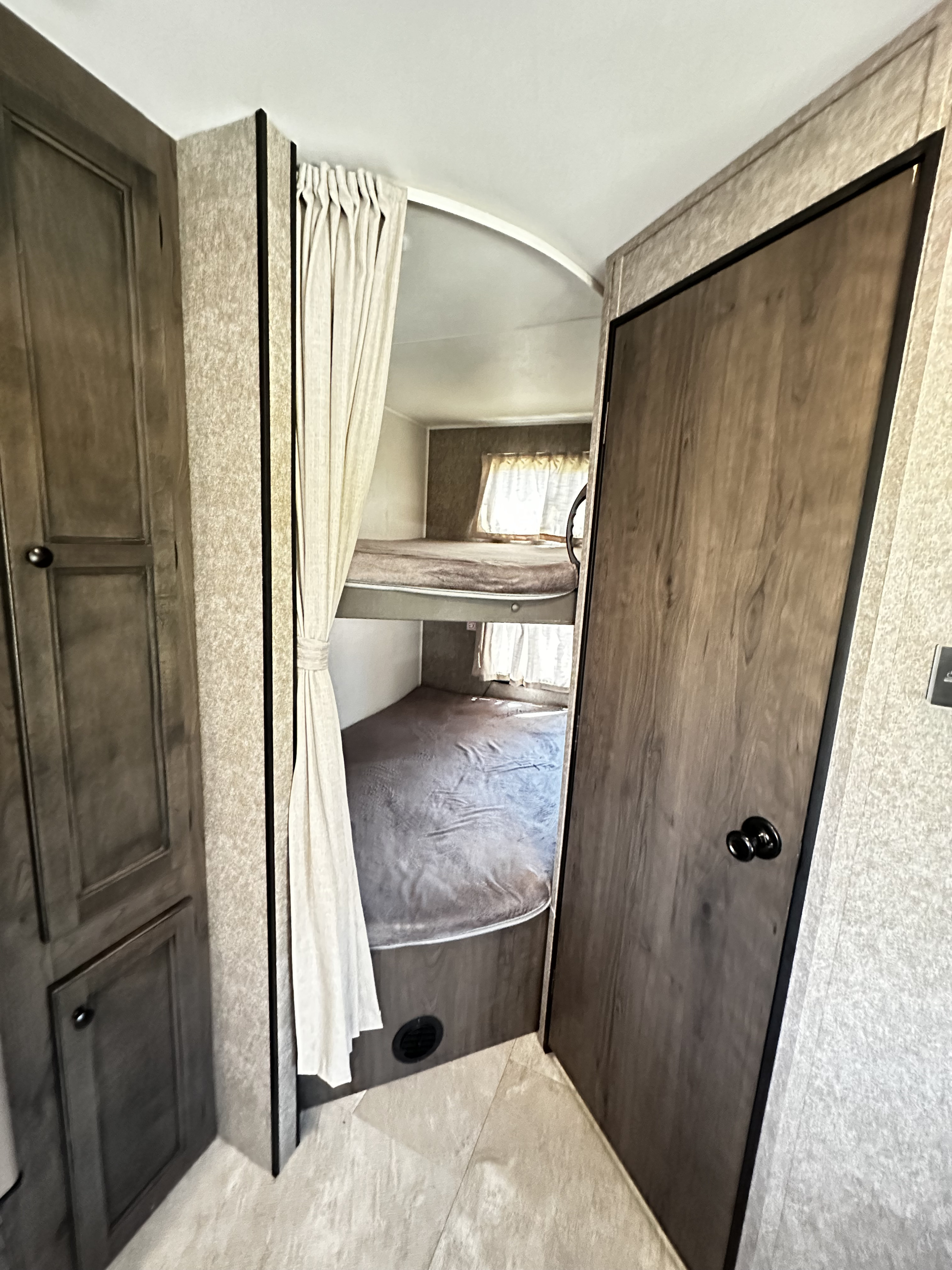 RV rental interior: Cozy bunk beds with grey bedding are visible behind a tied-back privacy curtain. Wooden cabinets flank the entry.. Starcraft Autumn Ridge 2021