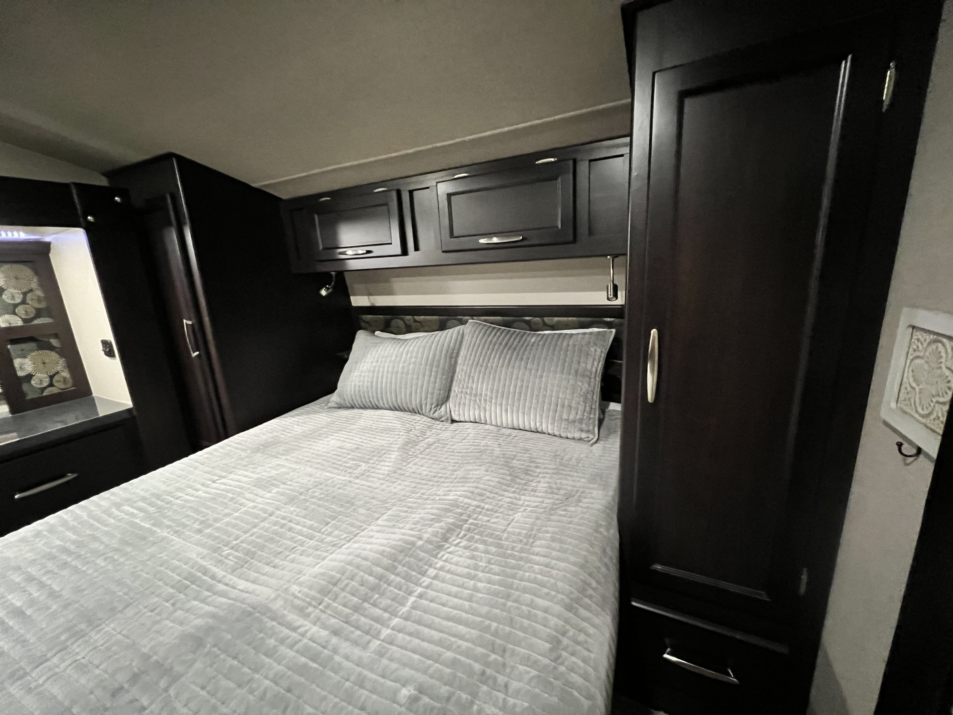 Compact RV rental bedroom with a comfortable grey bed, dark wood overhead cabinets, and built-in side storage.. Genesis Supreme Rv Genesis Supreme 2017