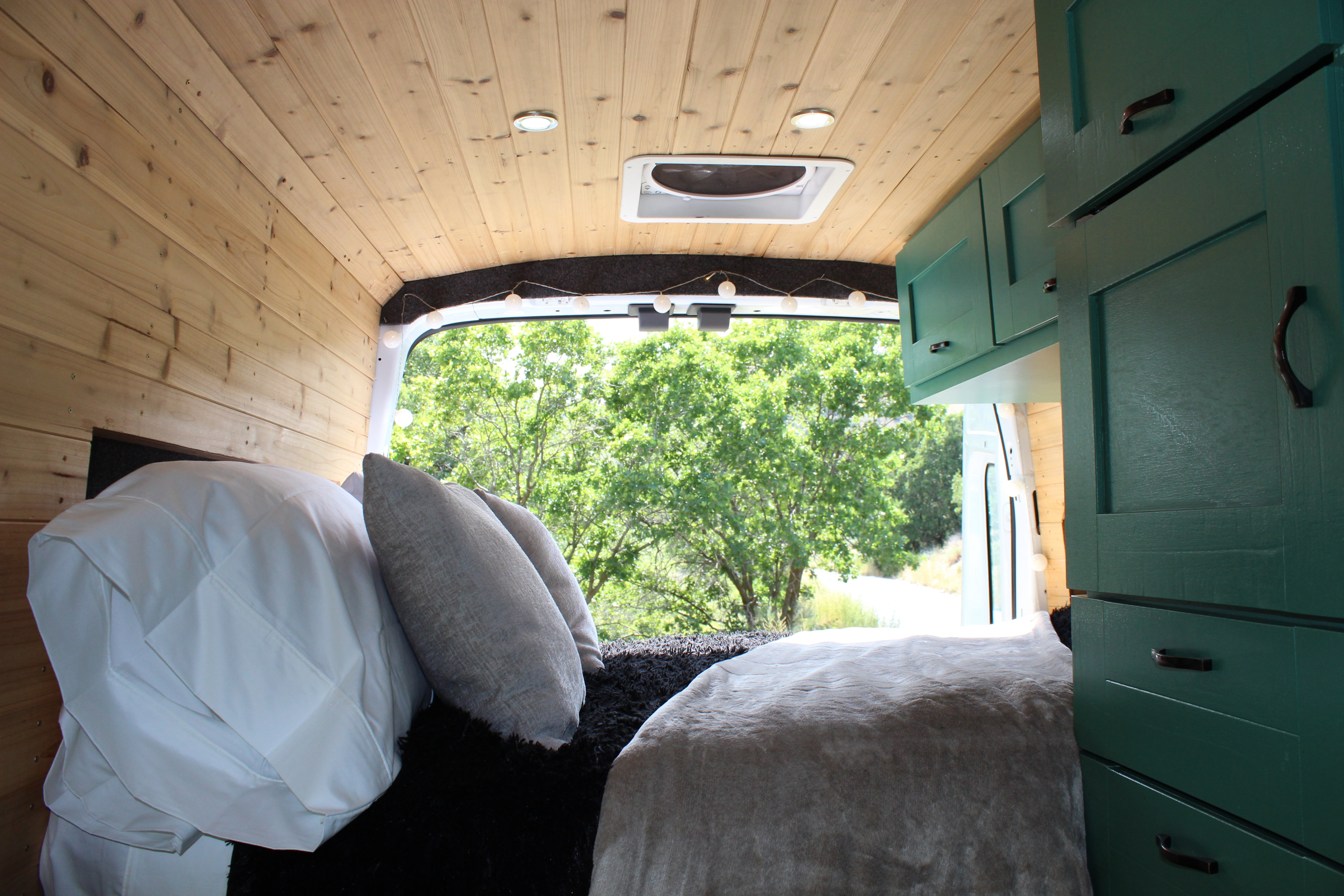 This RV rental interior features a cozy bed with pillows, wood-paneled walls/ceiling, green cabinets, and a serene outdoor view.. Ford Transit 2017
