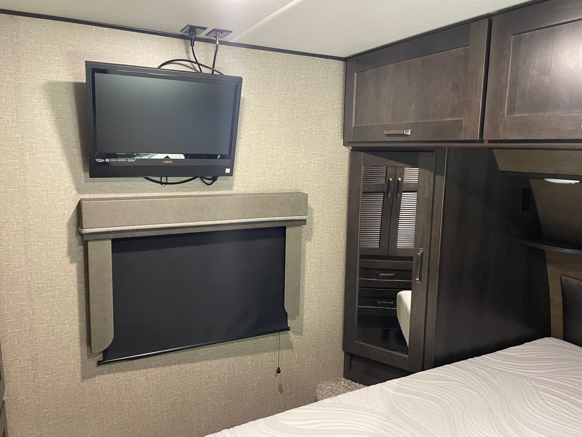 RV rental bedroom: TV, dark wood cabinets, and a white bed. A blackout shade covers the window on textured walls.. Grand Design Reflection 2022