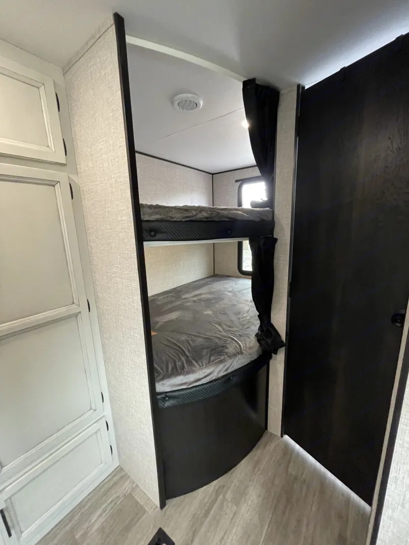 Inside an RV rental: a compact sleeping area with two neatly made bunk beds, privacy curtains, and light-colored cabinetry.. Jayco Jay Flight Baja Edition 2023