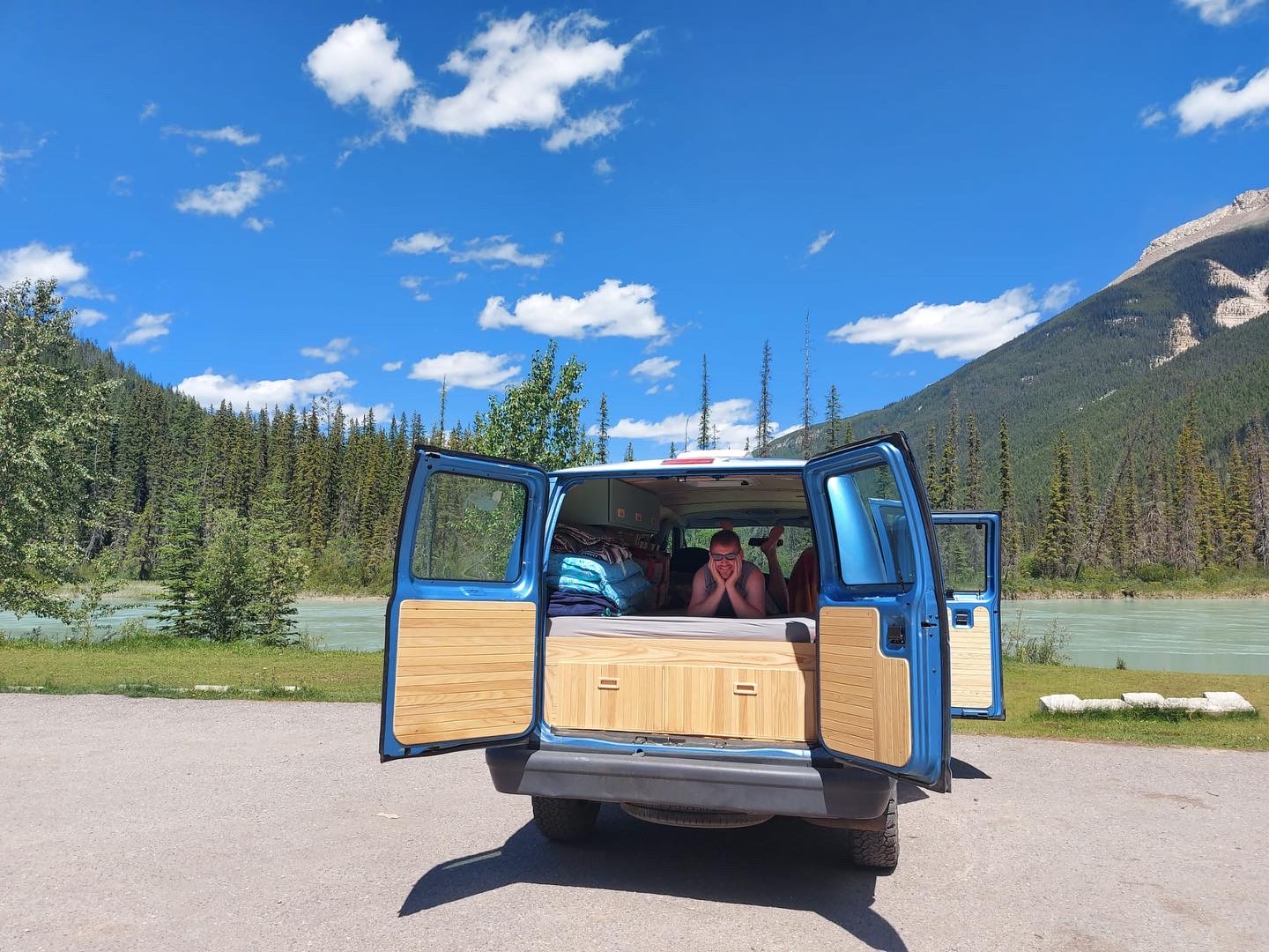 RV rental adventure: a blue campervan opens to a smiling person inside, overlooking a serene river, forest, and mountains.. Ford Econoline 2006