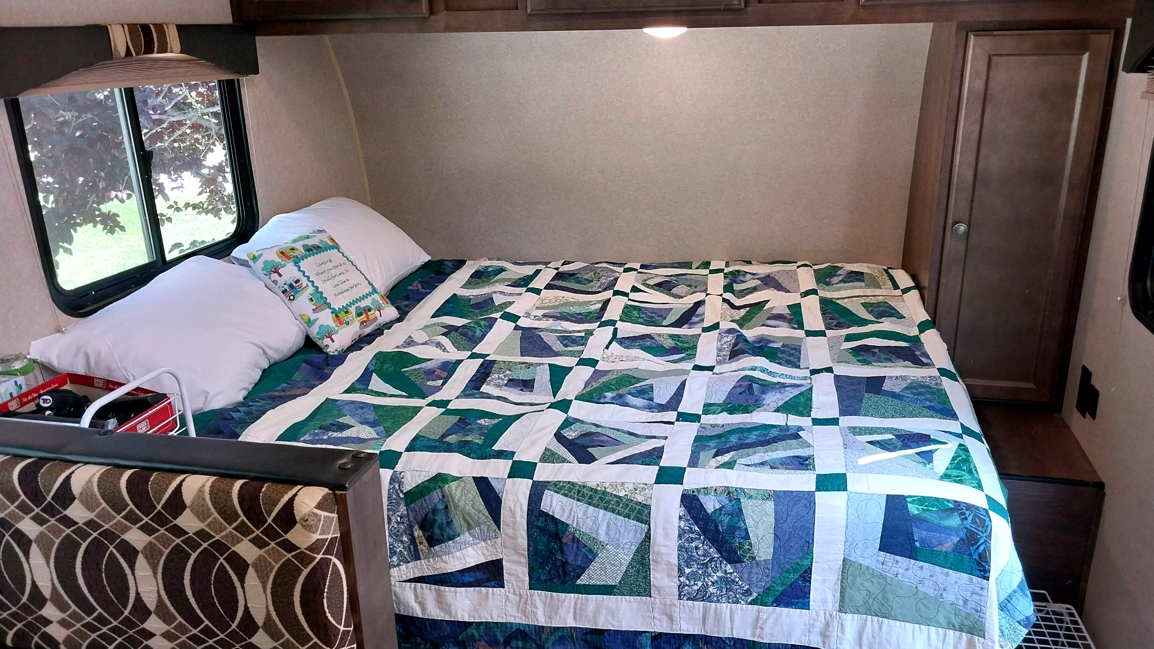 Inside an RV rental: a bed with a colorful patchwork quilt, white pillows, and a window showing lush trees.. Sunset Park RV Sun Lite 2018