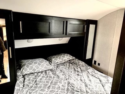 RV rental bedroom with gray patterned bed, dark overhead cabinets with lights, and a mirrored wardrobe for comfortable travel.. Forest River Cherokee Grey Wolf 2022
