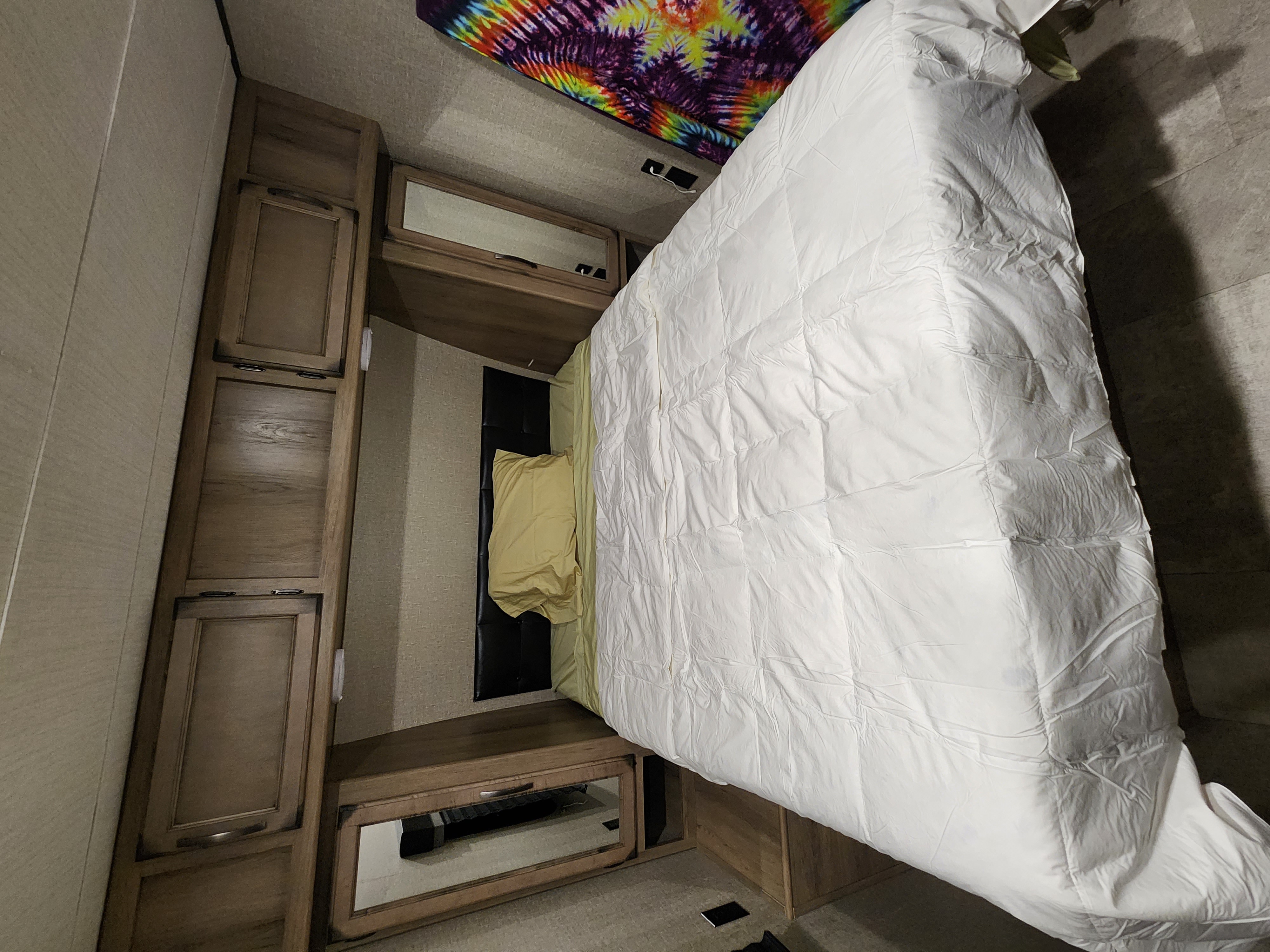 Cozy RV rental bedroom: bed with white comforter, yellow pillow, colorful tapestry, and wood cabinets for storage.. Grand Design Transcend 2019