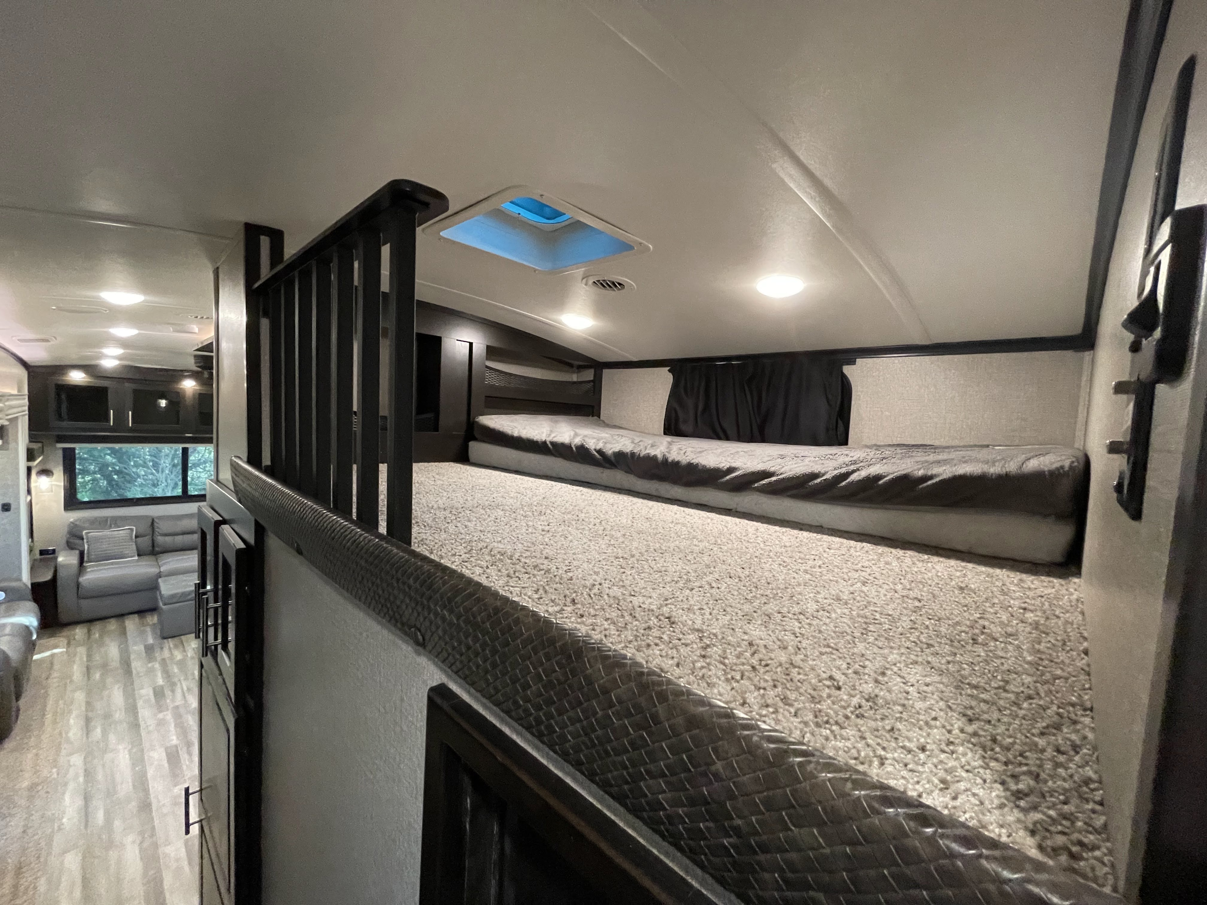 Modern RV rental interior showing a carpeted loft bunk with skylight, overlooking a bright living area with a gray sofa.. Jayco North Point 2020