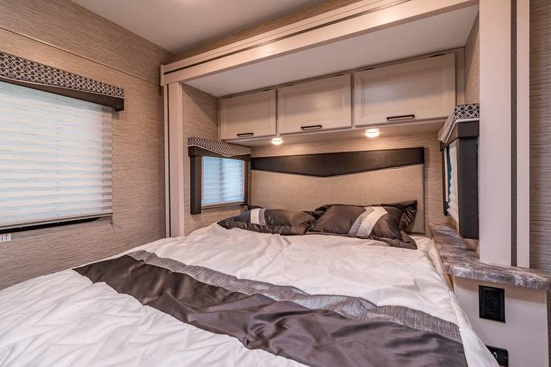 Spacious RV rental bedroom featuring a comfortable bed, overhead storage, and two windows with blinds, ideal for road trip comfort.. Thor Motor Coach Chateau 2023