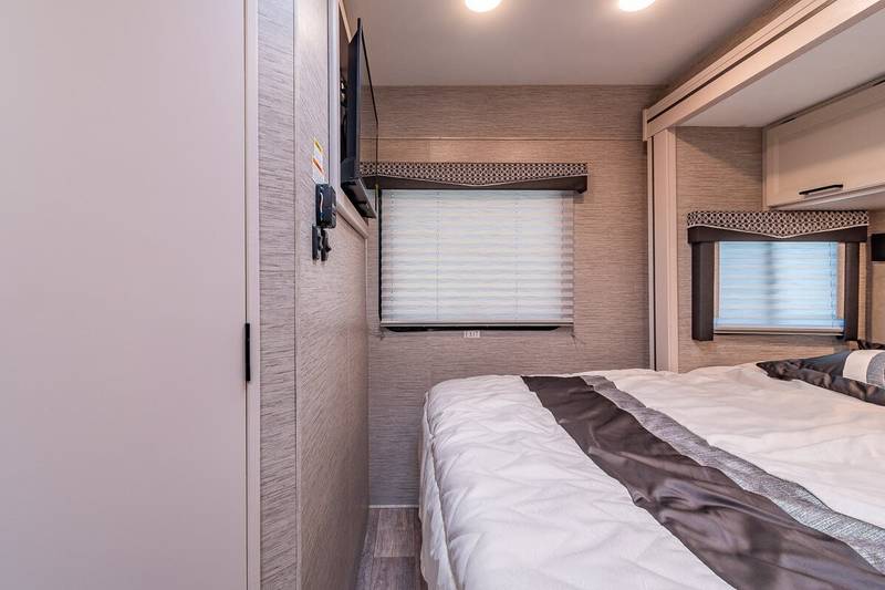 An RV rental's compact bedroom features a comfortable bed, wall-mounted TV, and blinds on two windows.. Thor Motor Coach Chateau 2023