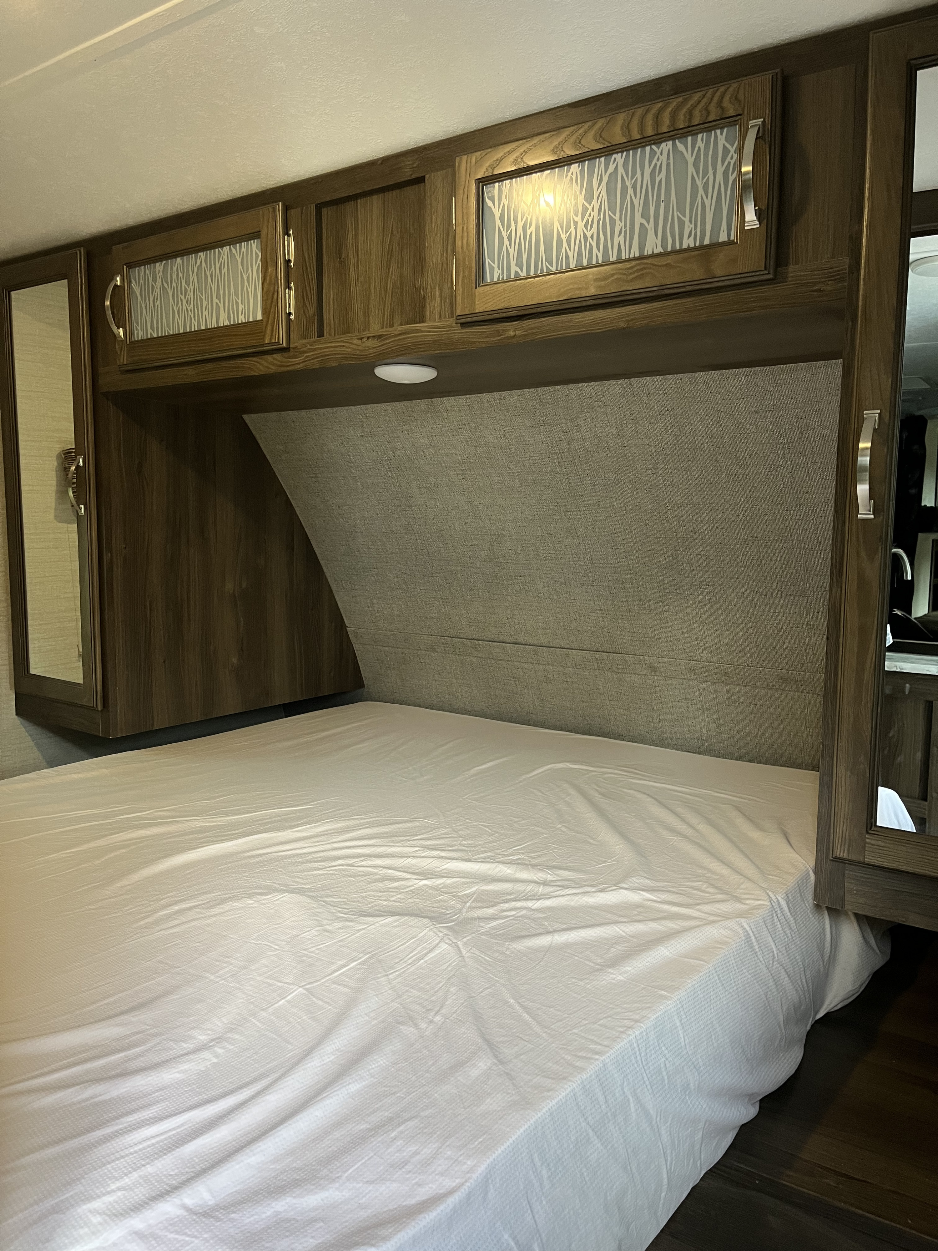 An RV rental's cozy bedroom features a bed with white sheets, wood overhead cabinets, and a textured headboard.. Keystone RV Bullet Ultra Lite 2019
