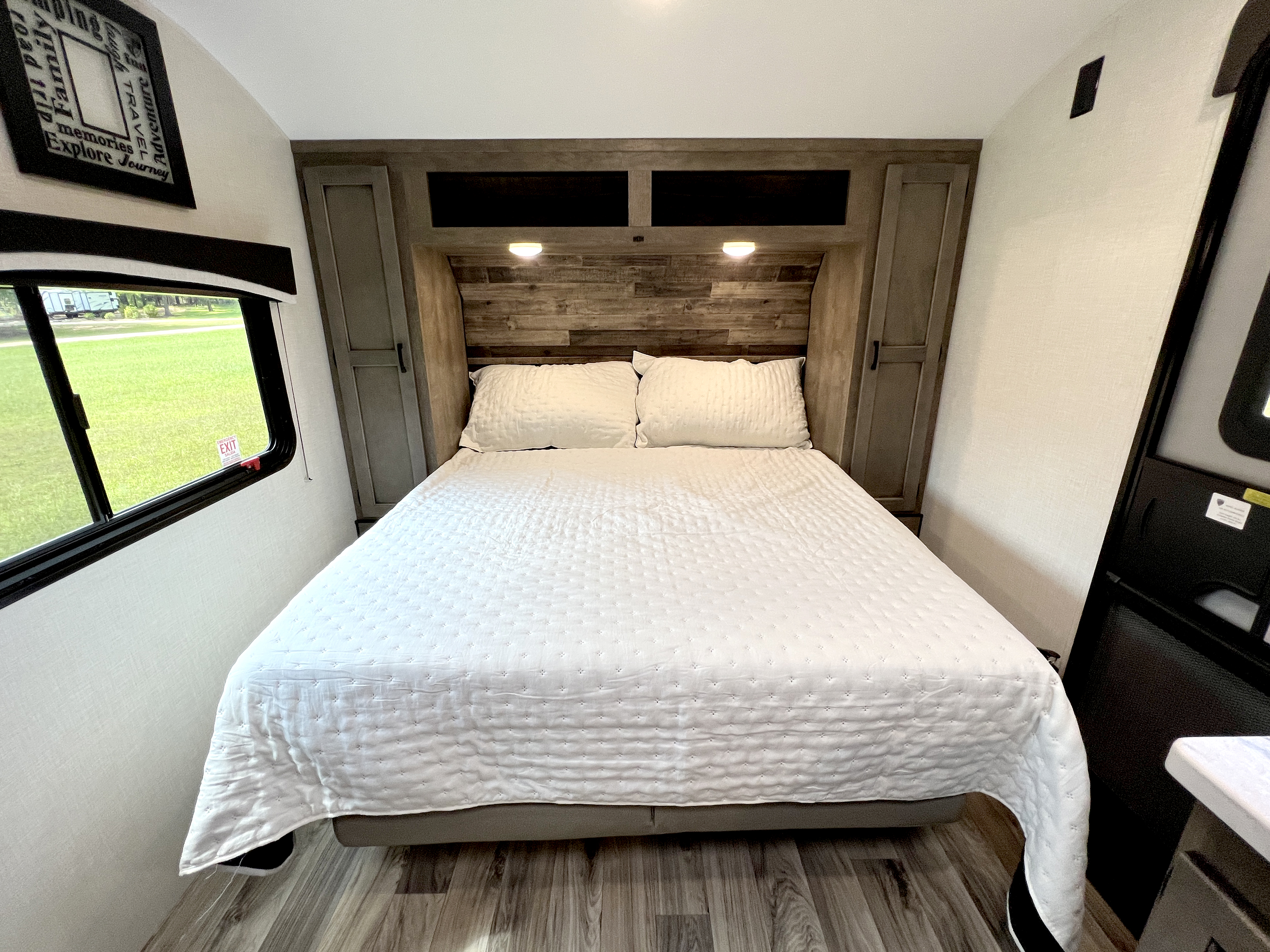 Well-lit RV rental bedroom features a comfy bed, wood headboard, cabinets, and a window with green views.. Venture Rv Sonic 2023