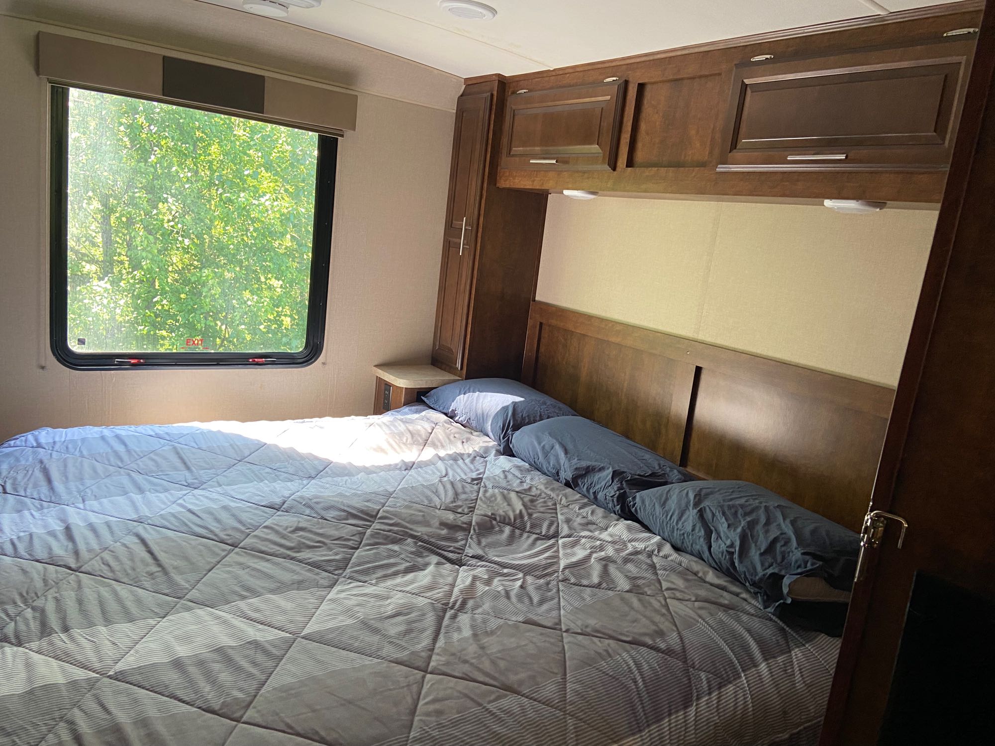 Cozy RV rental bedroom with a comfortable bed, wooden cabinets, and a large window offering a lush green tree view.. Forest River Fr3 2017