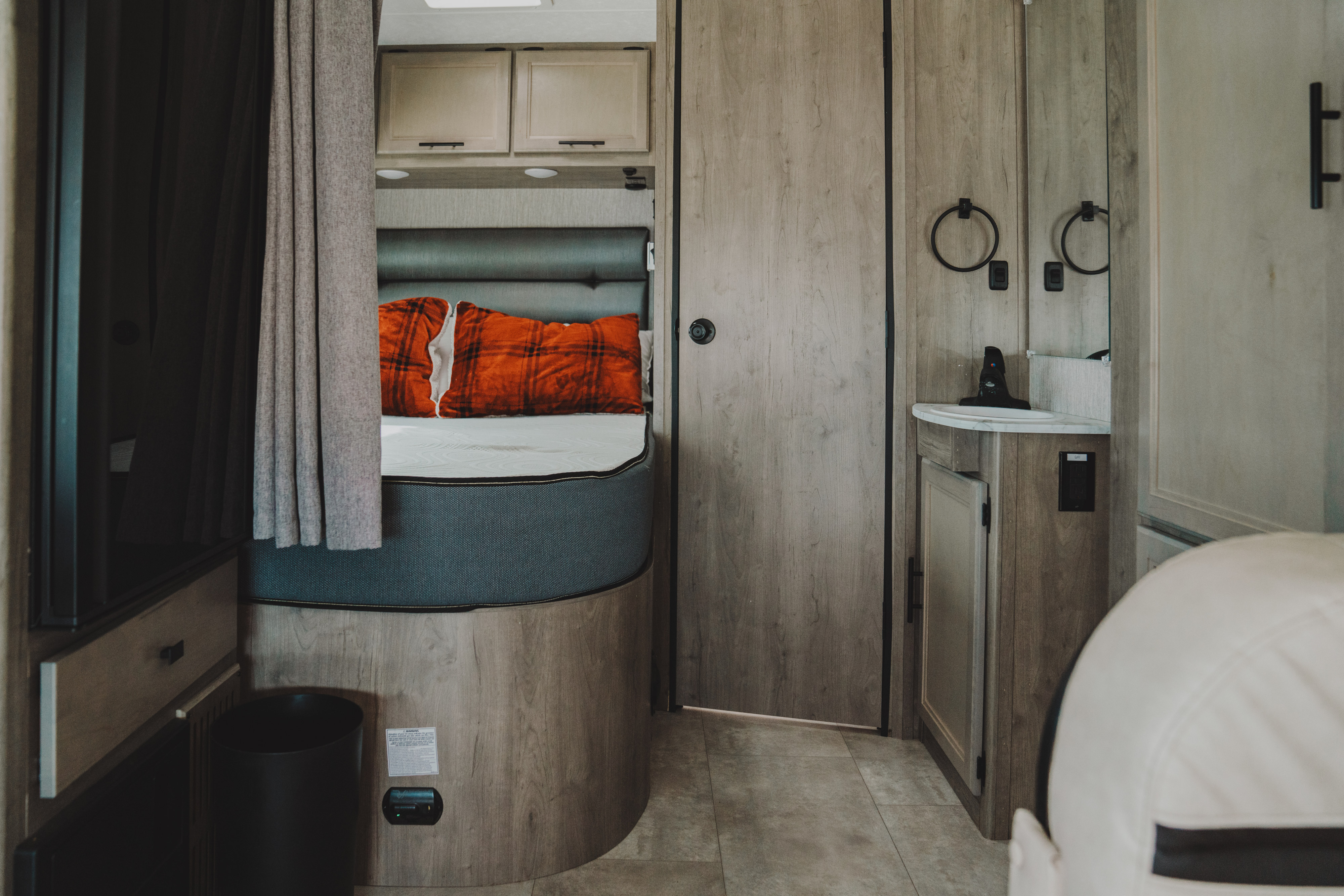 This rv rental features a cozy bedroom with orange plaid pillows, overhead cabinets, and a functional vanity/sink area.. Coachmen Prism 2021