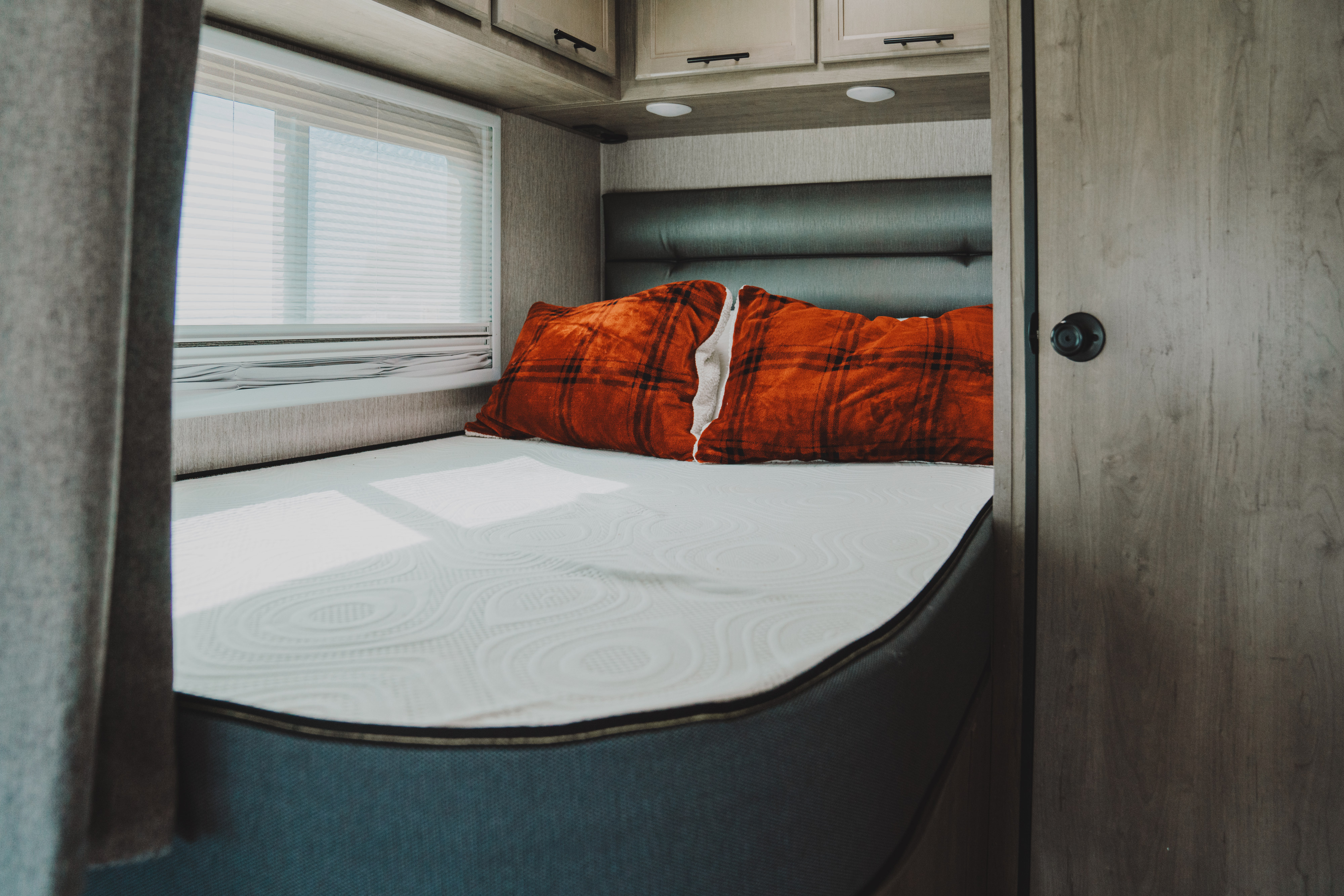 Cozy RV rental bedroom features a comfortable bed, plaid pillows, window blinds, and overhead storage for your adventure.. Coachmen Prism 2021