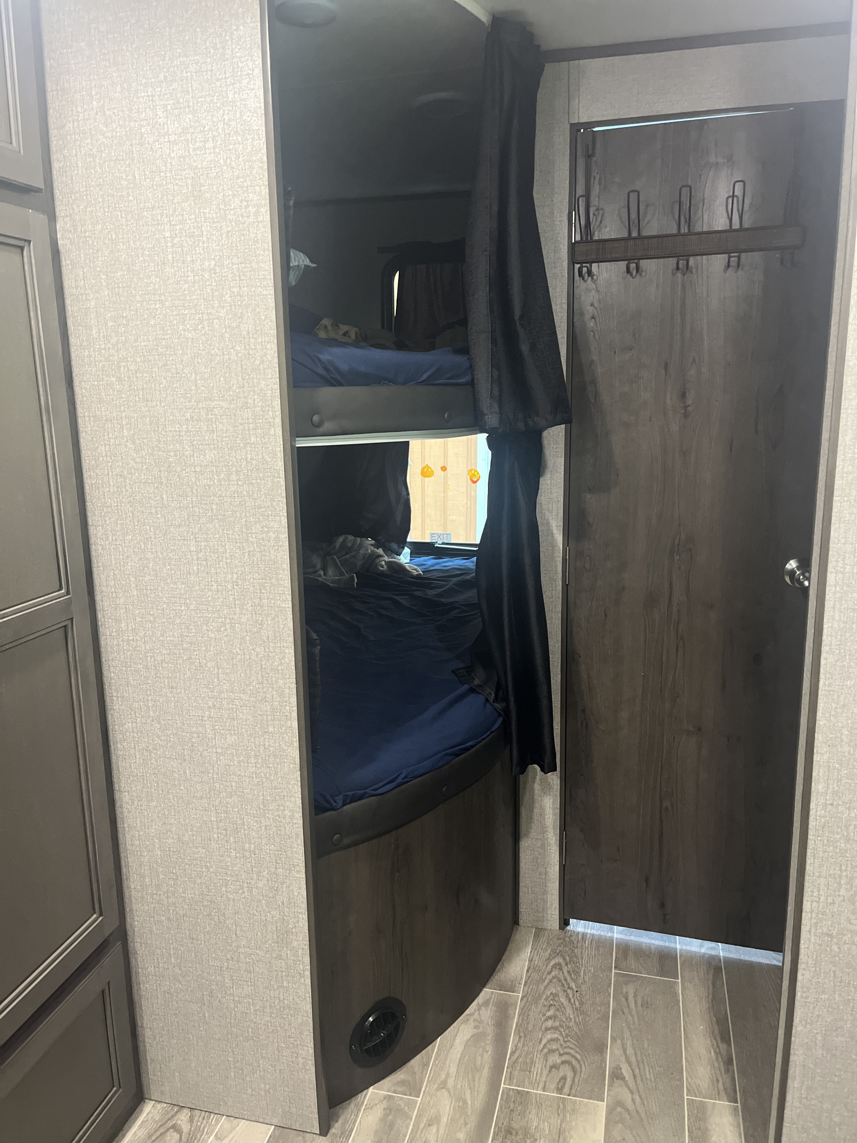 An RV rental features two bunk beds with dark privacy curtains, a window, floor vent, and a door with coat hooks.. Jayco Jay Flight SLX 2021