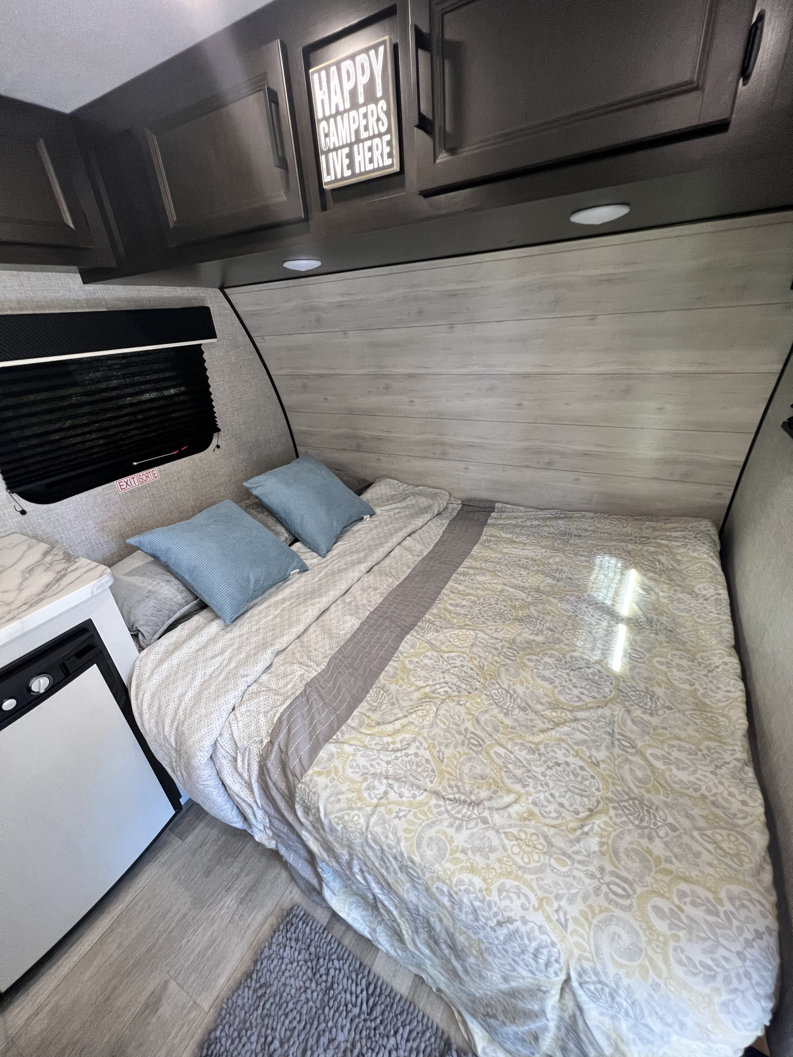 Cozy RV rental bedroom: bed with blue pillows, overhead cabinets, small fridge, and a "Happy Campers" sign.. Jayco Jay Flight SLX Baja Edition 2023