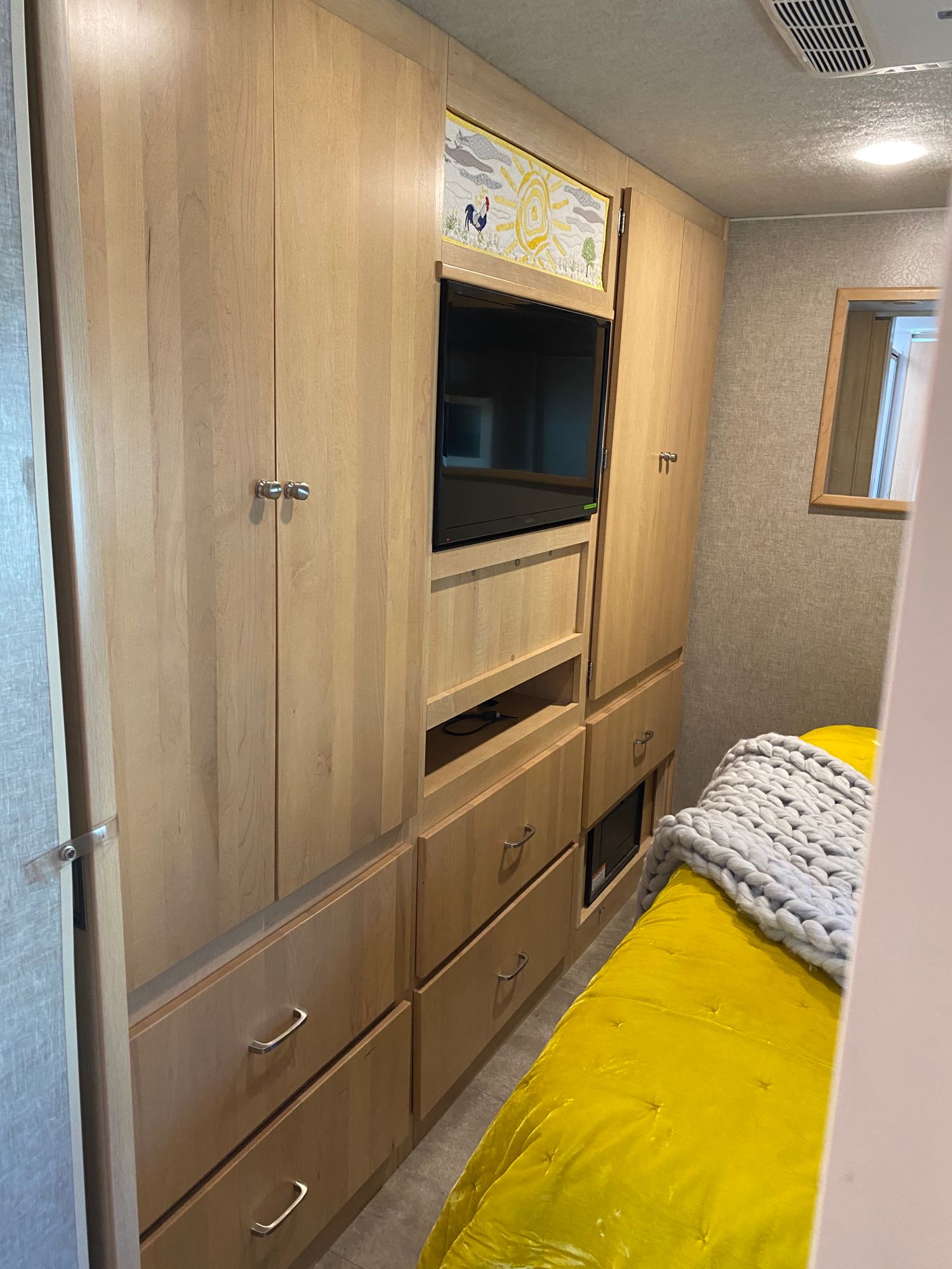 RV rental interior: ample light wood storage, built-in TV, and a cozy bed with bright yellow bedding and a grey blanket.. Winnebago Brave 2016