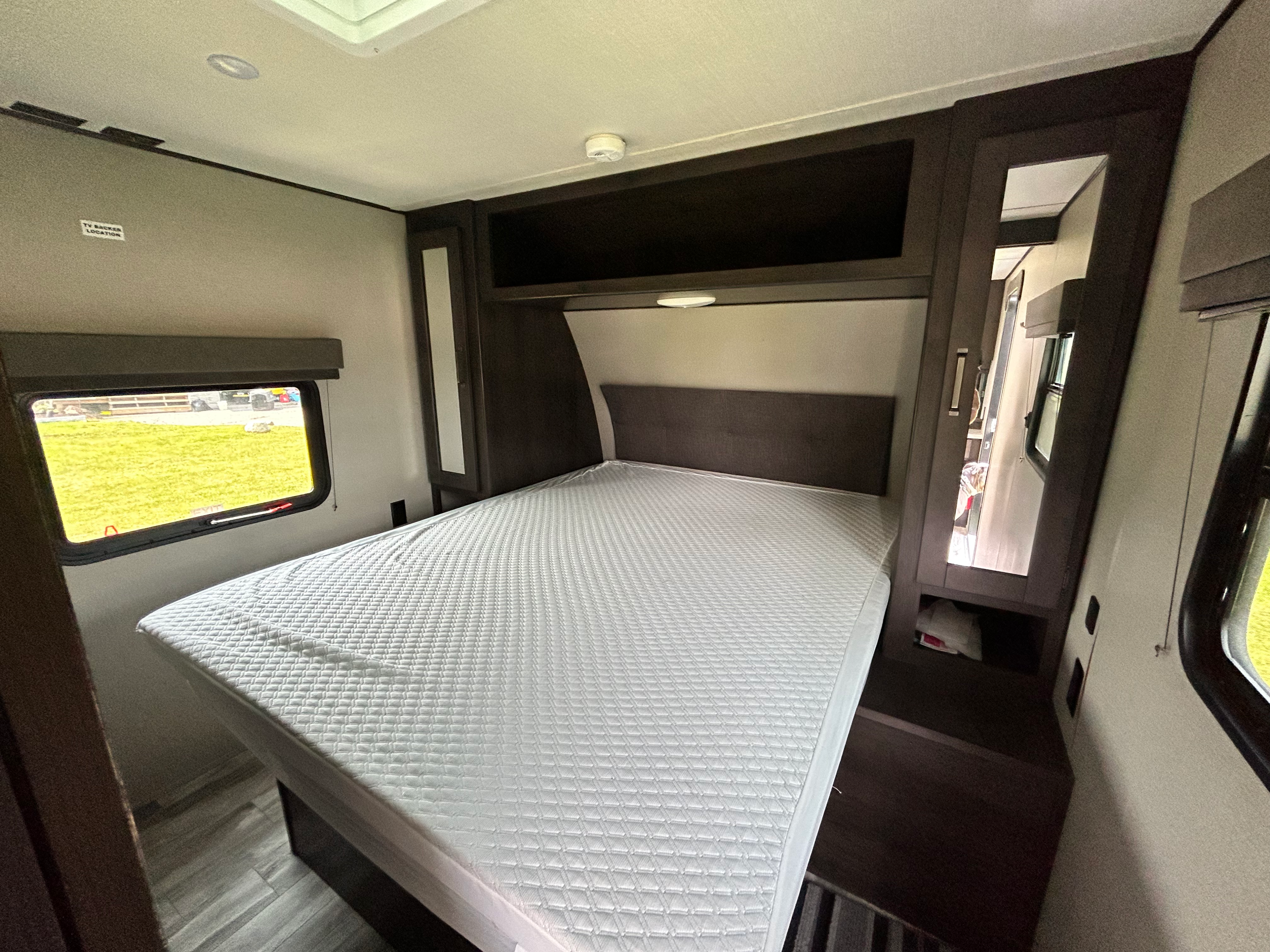 Cozy RV rental bedroom showcasing a bed with a white quilted mattress, overhead storage, and a large window overlooking green grass.. Grand Design Transcend Xplor 2021