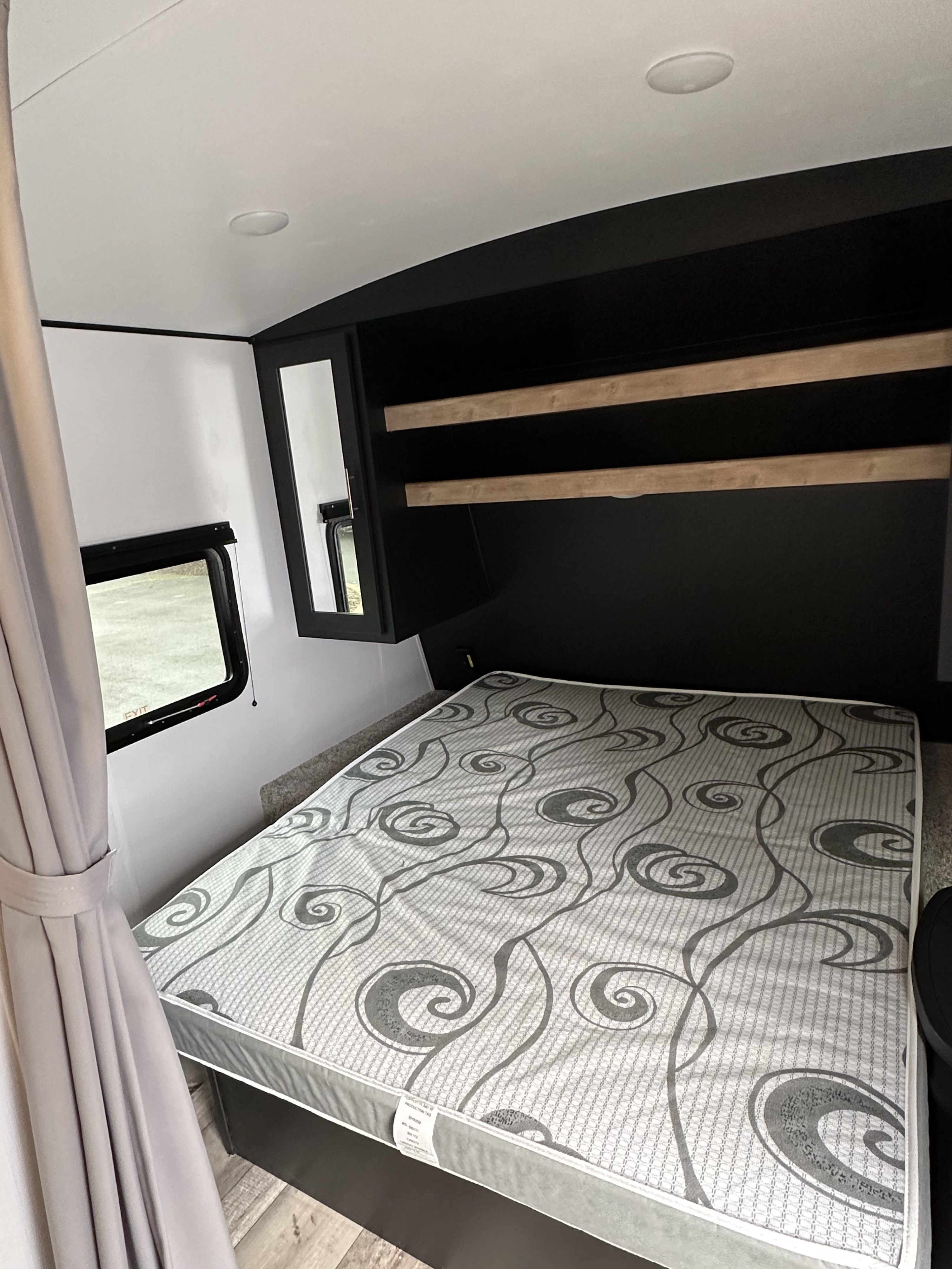 Cozy RV rental bedroom with a patterned mattress, black headboard with shelves, and a window with light-filtering curtains.. Keystone RV Springdale 2022