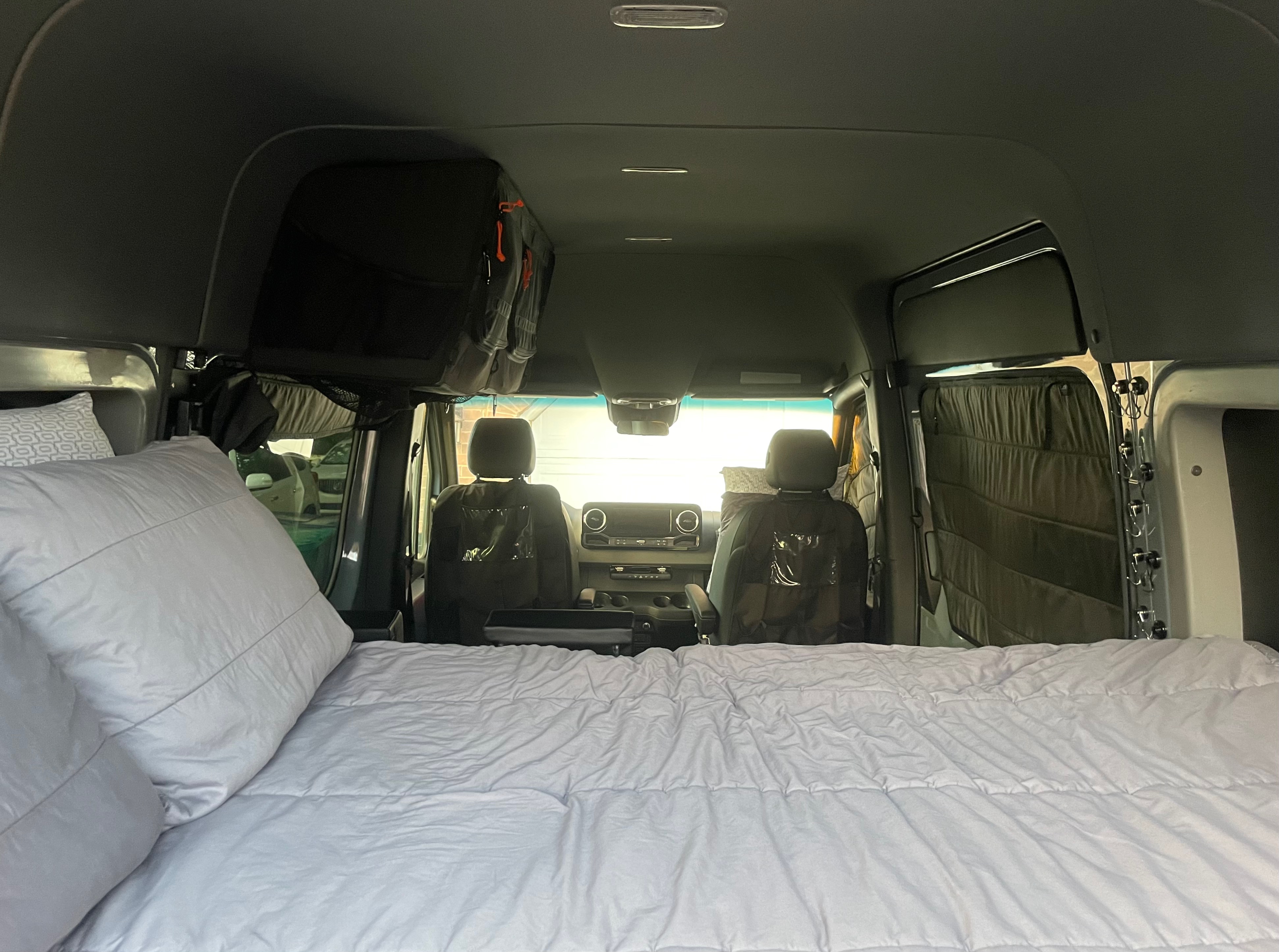 Inside an RV rental, a comfy bed fills the foreground, leading to the front cabin with seats and overhead storage.. Mercedes-Benz Sprinter 2019