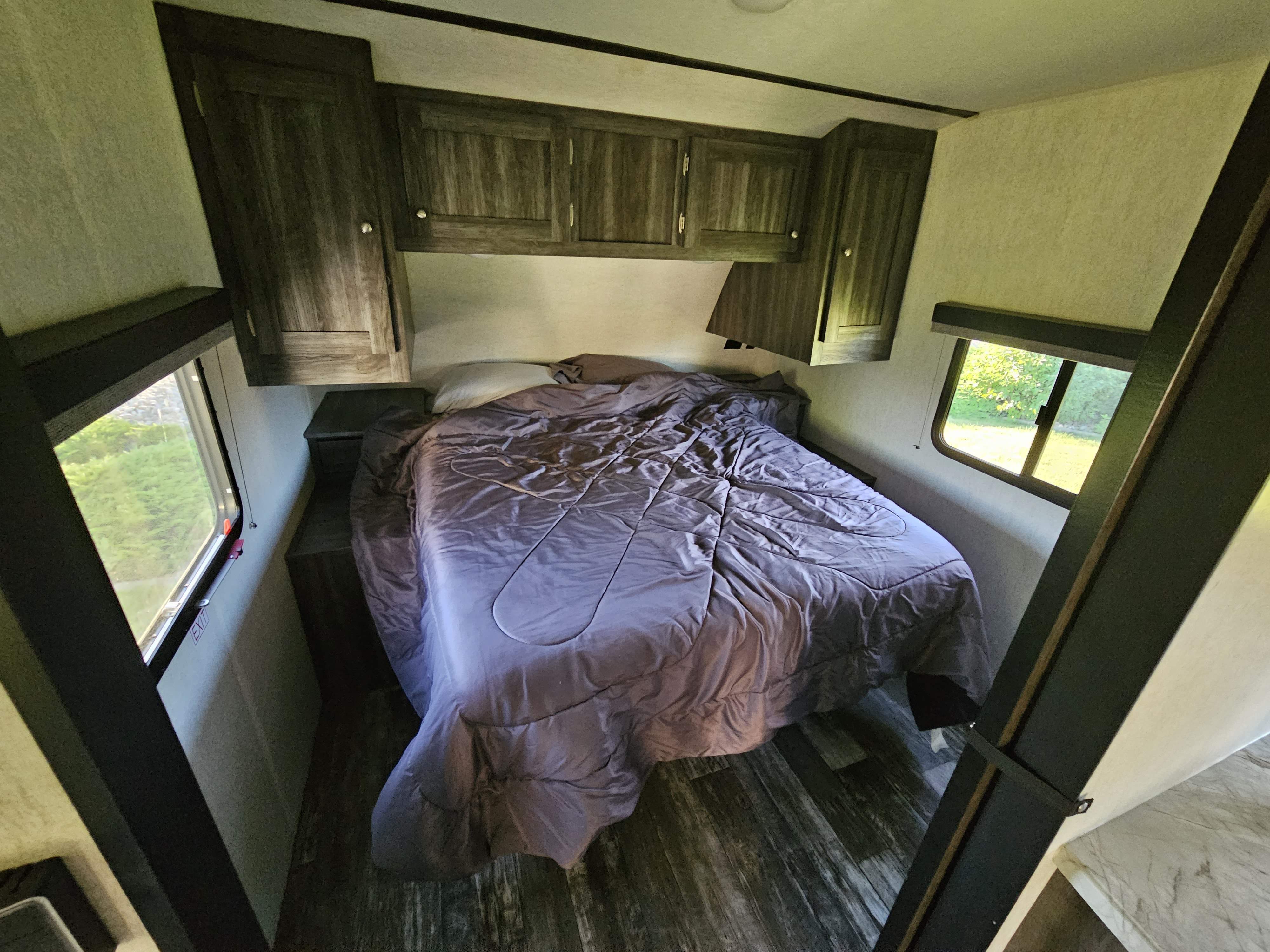 This cozy rv rental bedroom features a bed with a gray comforter, dark wood cabinets, and windows offering natural light and outdoor views.. Starcraft Super Lite 2019