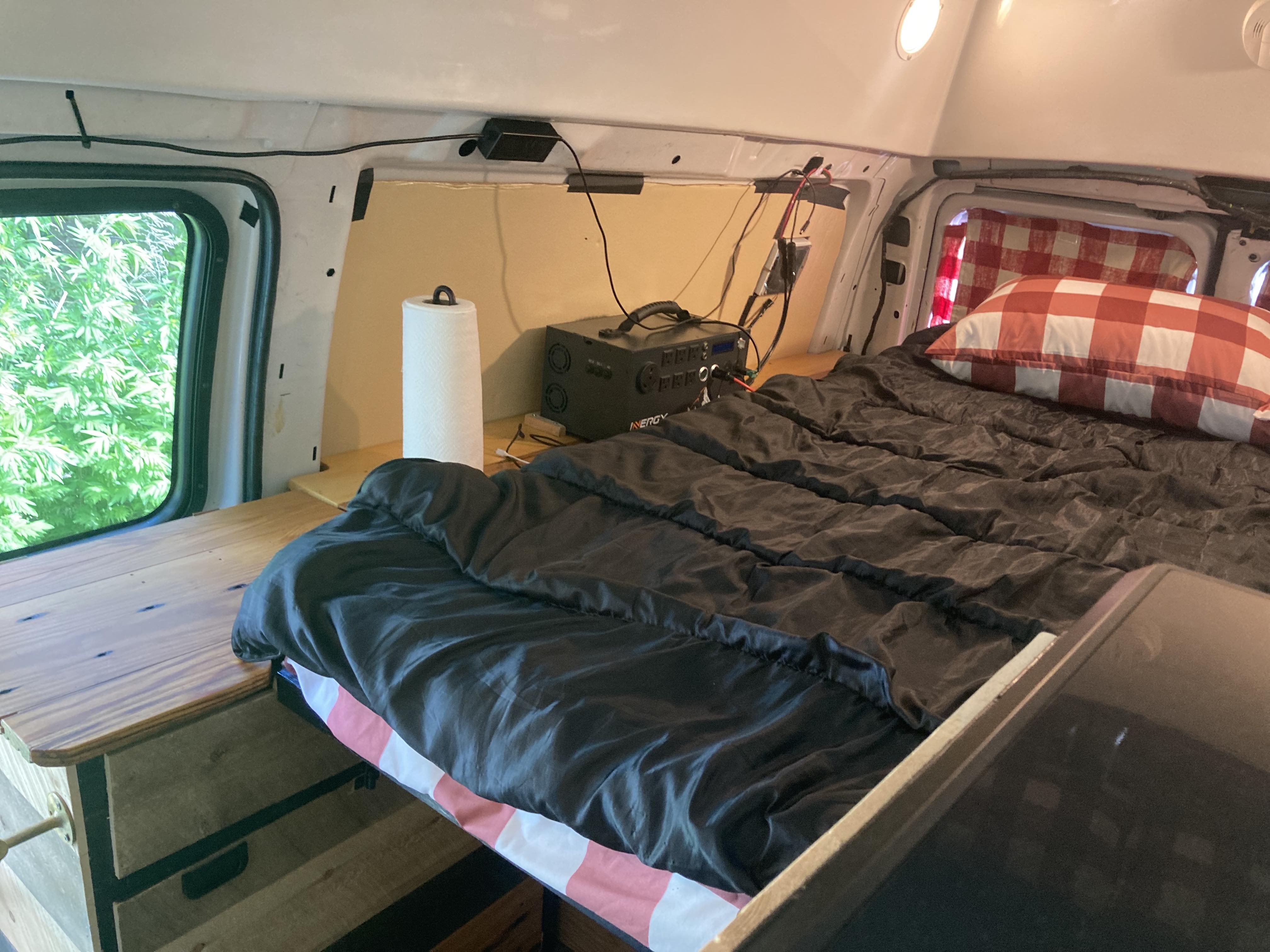 Cozy RV rental campervan interior: bed with checkered pillow, portable power station, and wooden table/shelf.. Ford E350 Box Van 2003