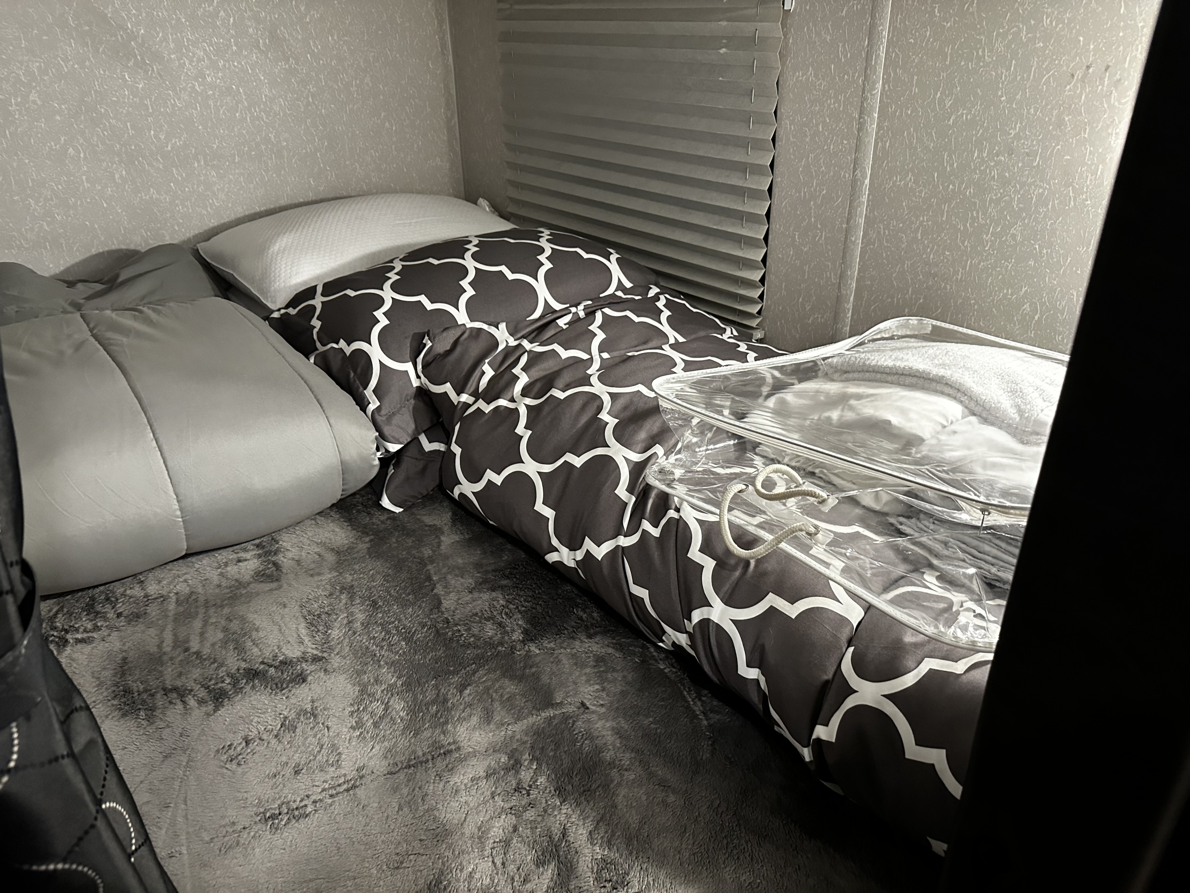 Cozy RV rental bed with patterned comforter, gray blanket, window blinds, and clear storage bag.. Forest River Cherokee Grey Wolf Black Label 2021