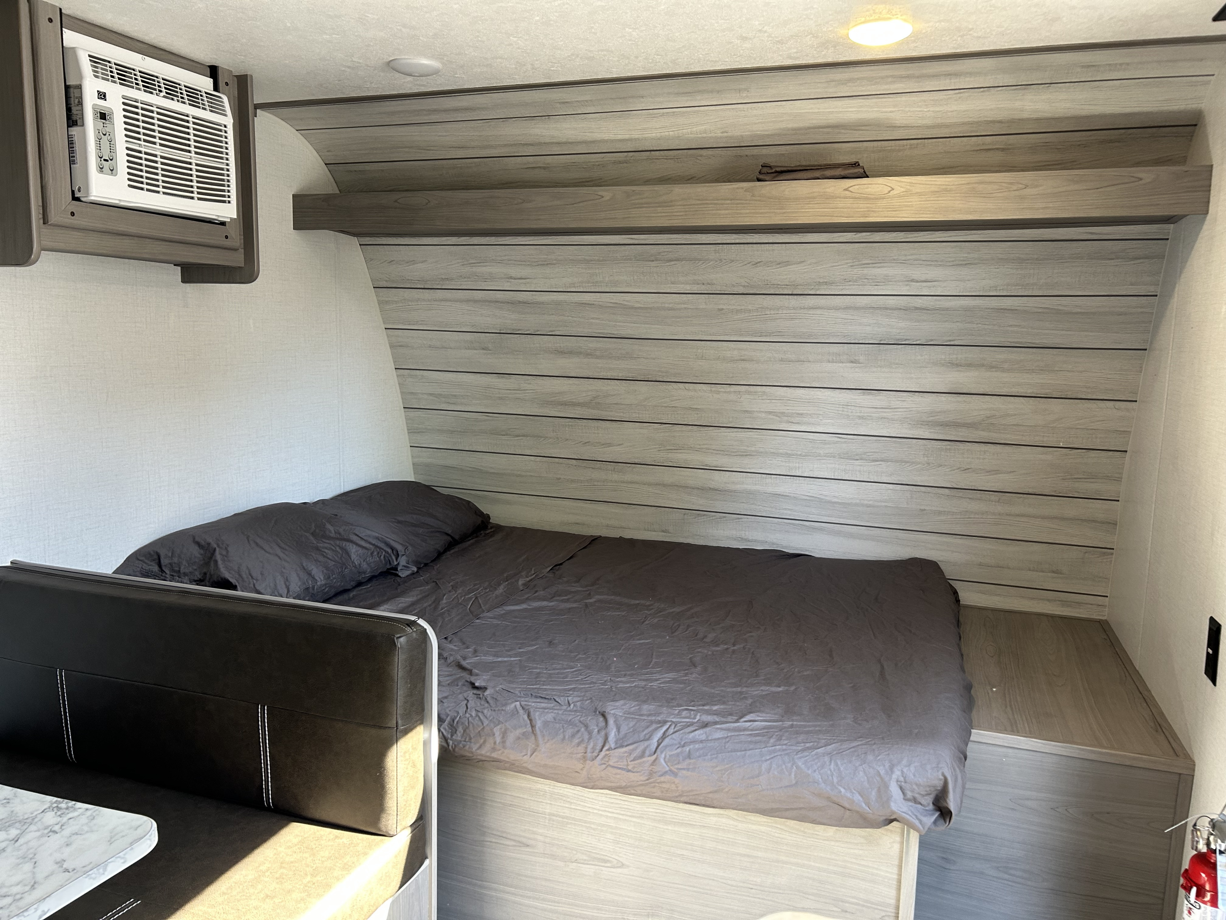 Inside an RV rental: a compact bedroom with a dark grey bed, overhead shelf, and AC unit near the dinette.. Dutchmen Coleman Lantern LT 2023