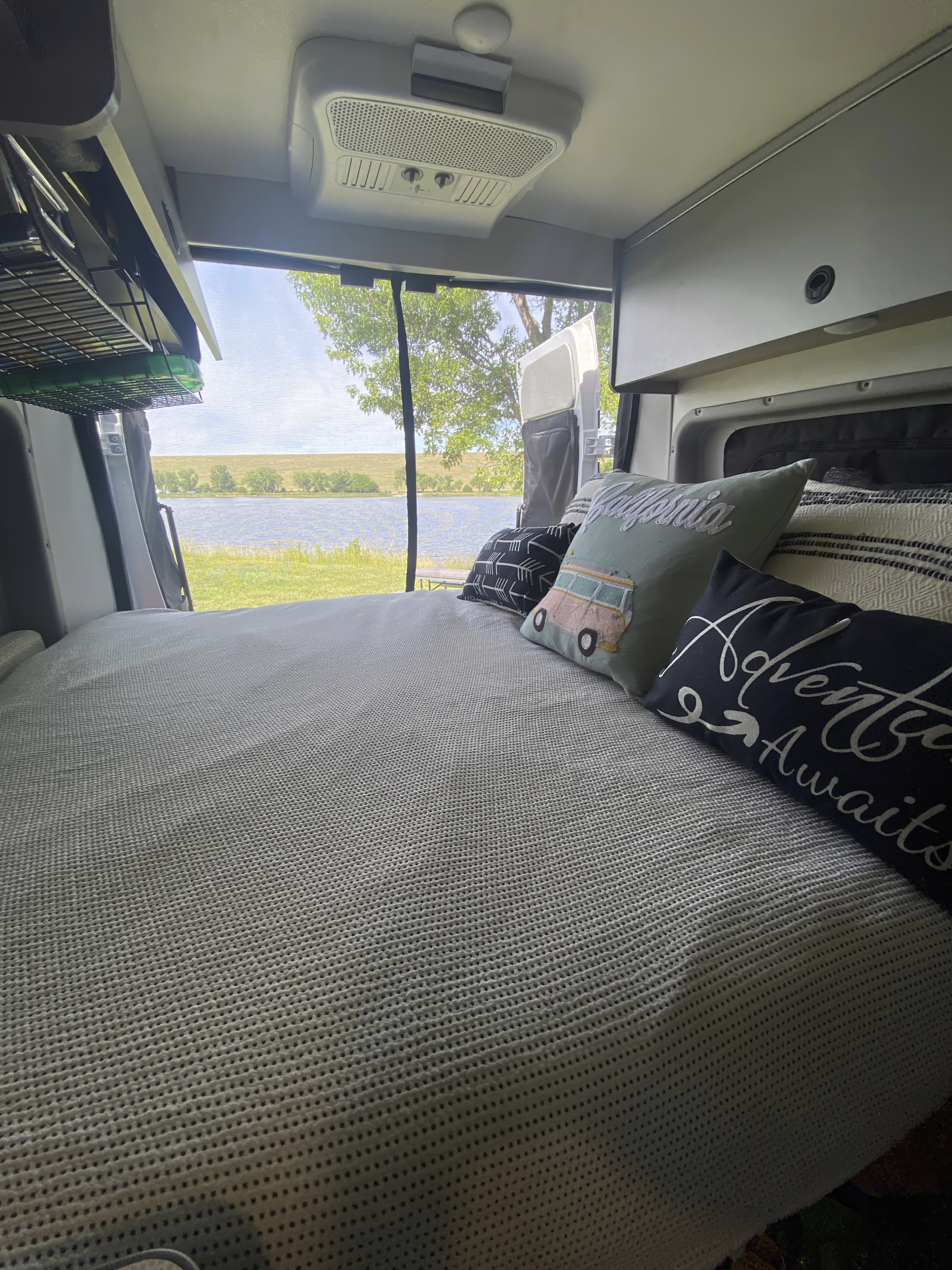 Inside an RV rental: a comfortable bed with travel-themed pillows overlooks a serene lake and green landscape.. Winnebago Solis 2021