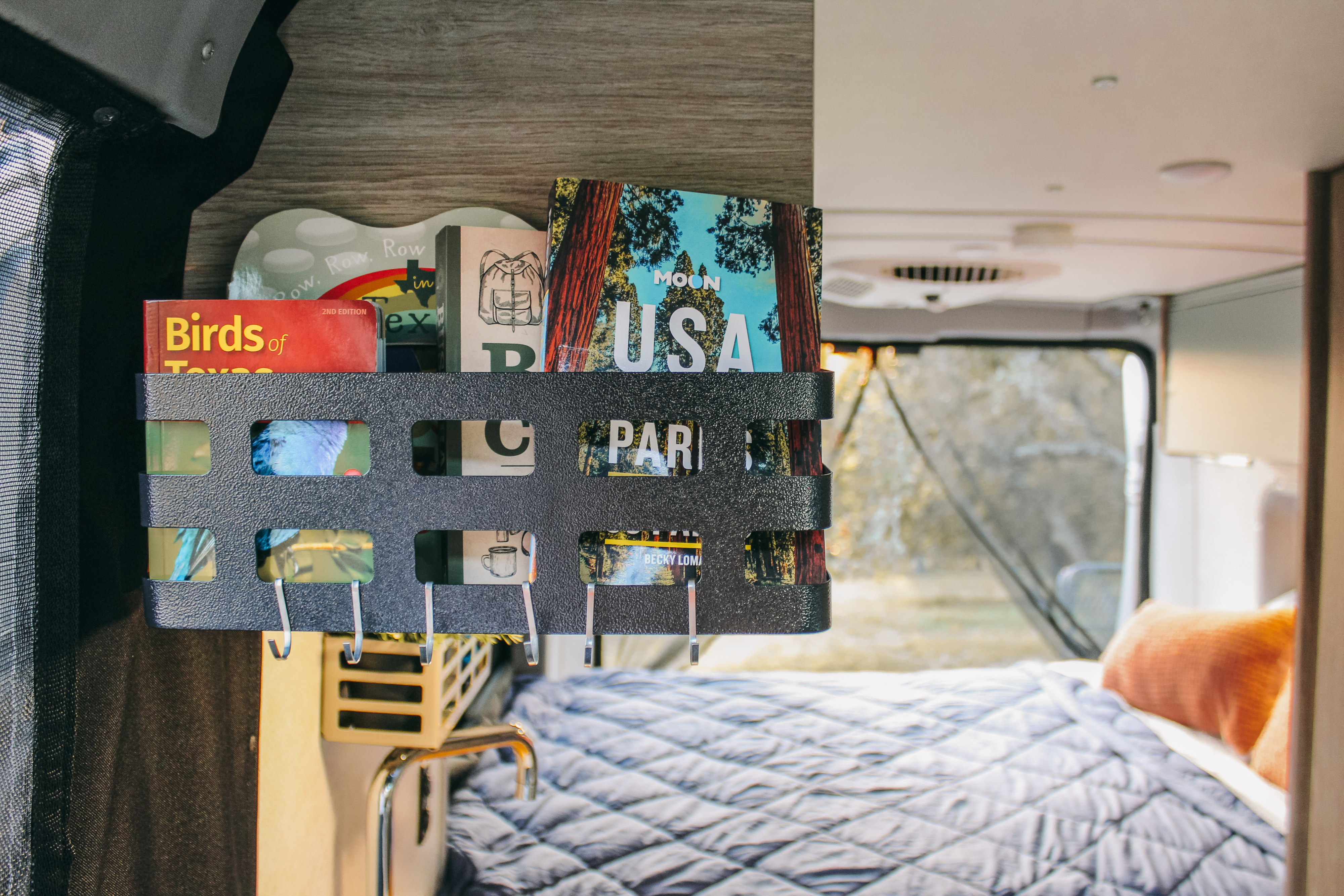 RV rental: Organized travel guides like "Moon USA" & "Birds of Texas" on a black wall rack, with a cozy bed inside.. Winnebago Solis 2023