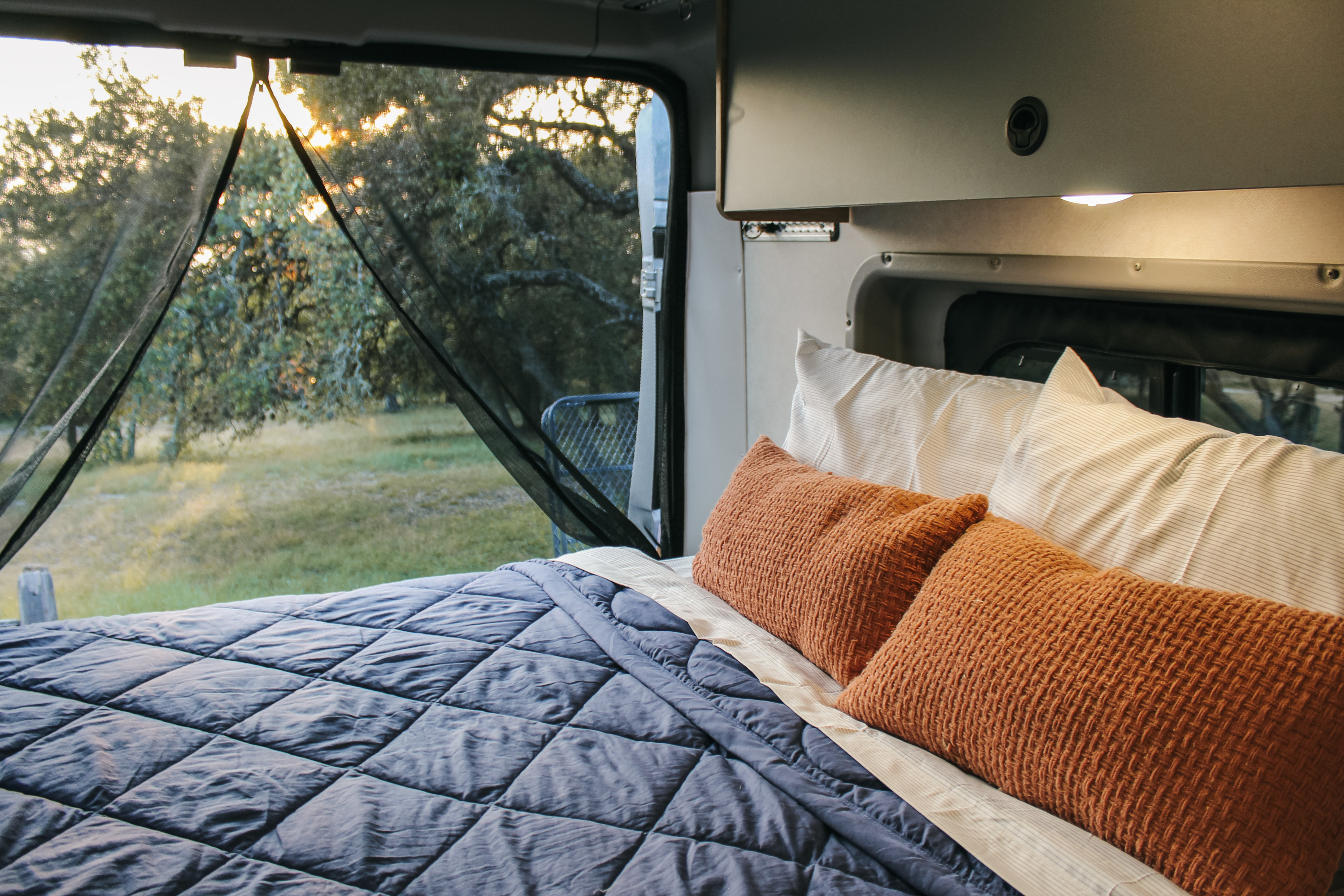 Cozy RV rental bedroom with blue comforter & orange pillows, offering serene nature views through a mesh screen.. Winnebago Solis 2023