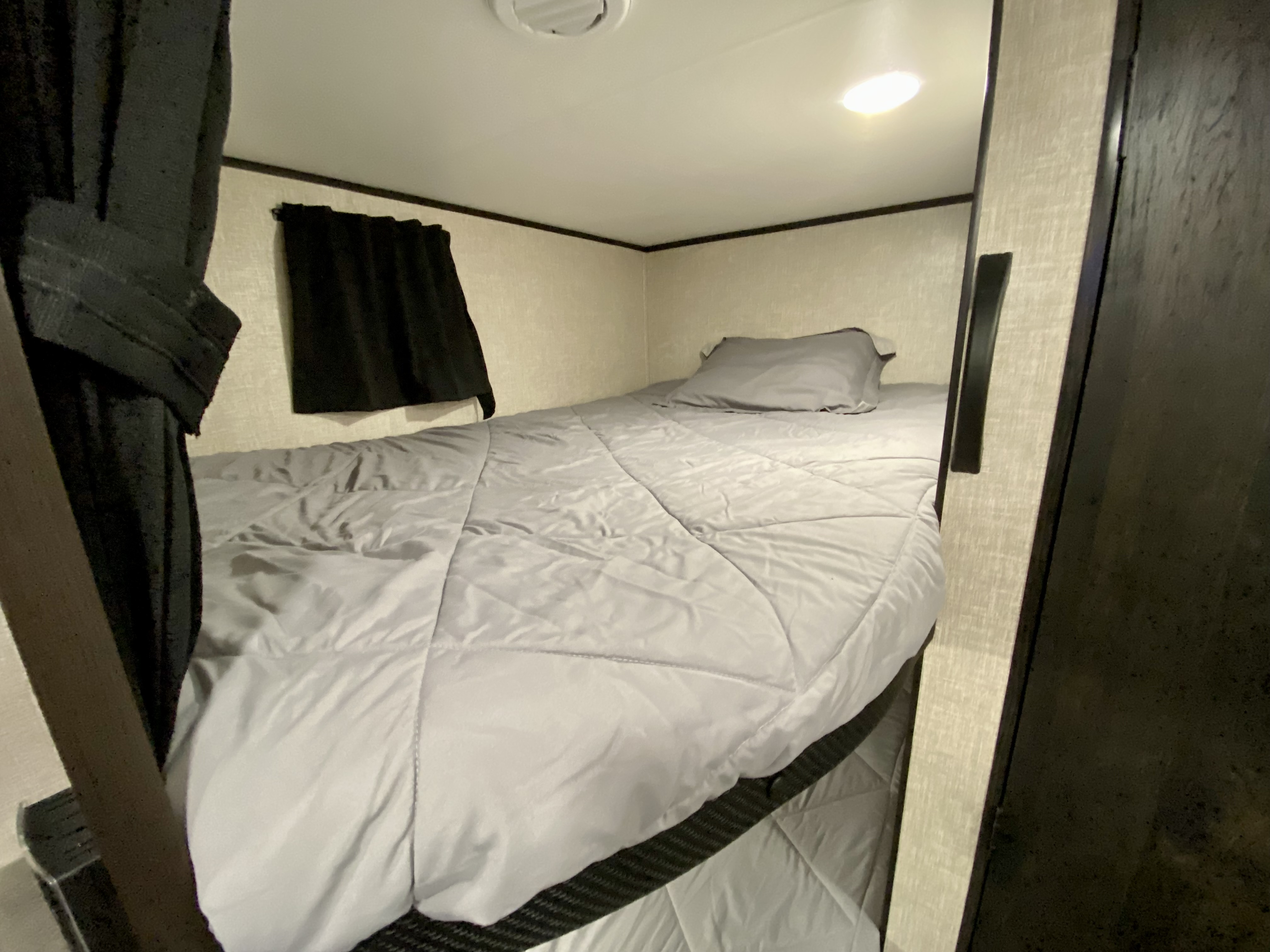 Compact RV rental sleeping area features a comfy grey bed, window with dark curtain, and ceiling light.. Jayco Jay Flight 2023