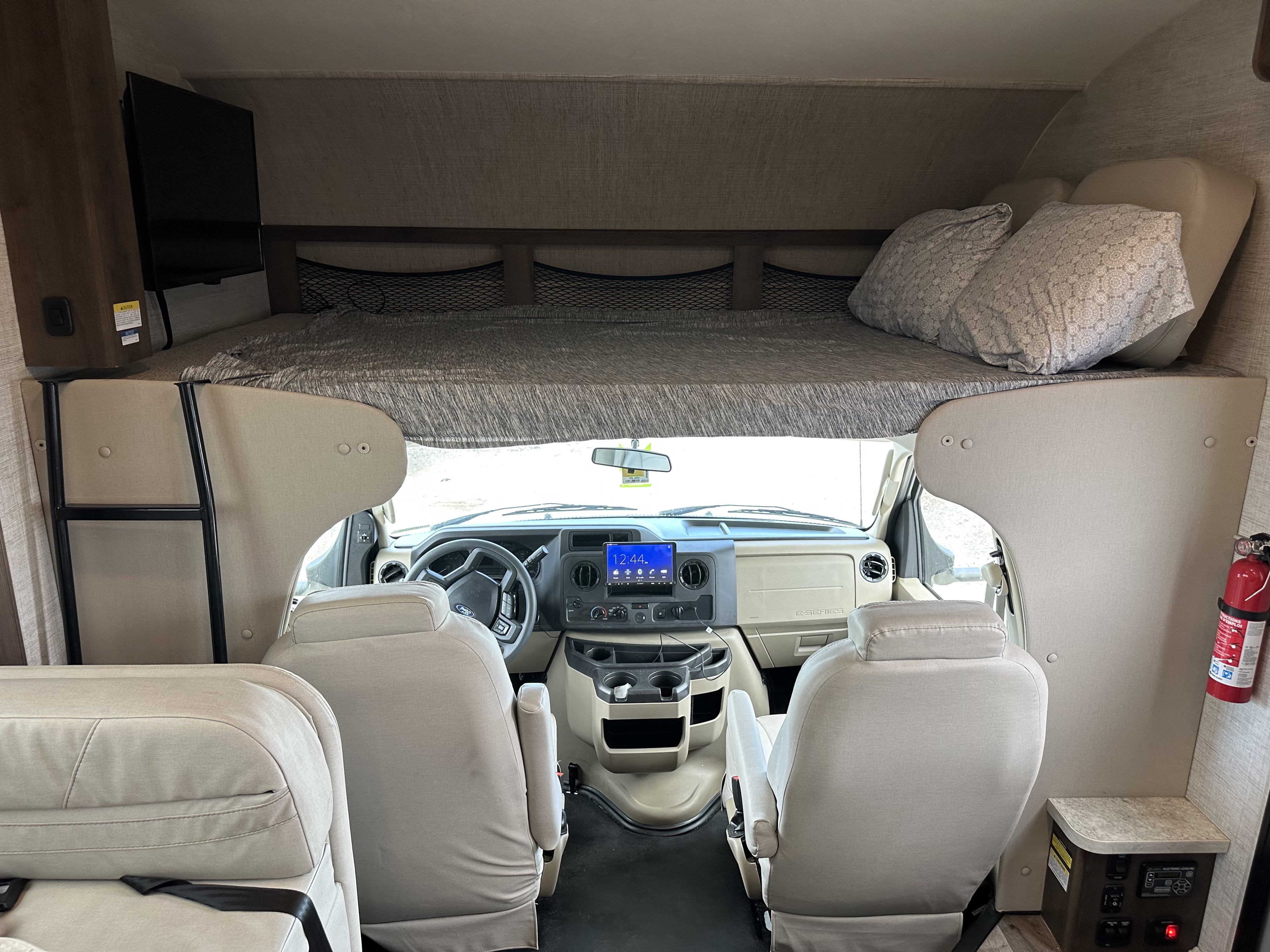 Inside an RV rental: cockpit with steering wheel & screen, front seats, and an overcab bunk bed with pillows, TV, and ladder.. Jayco Redhawk 2021