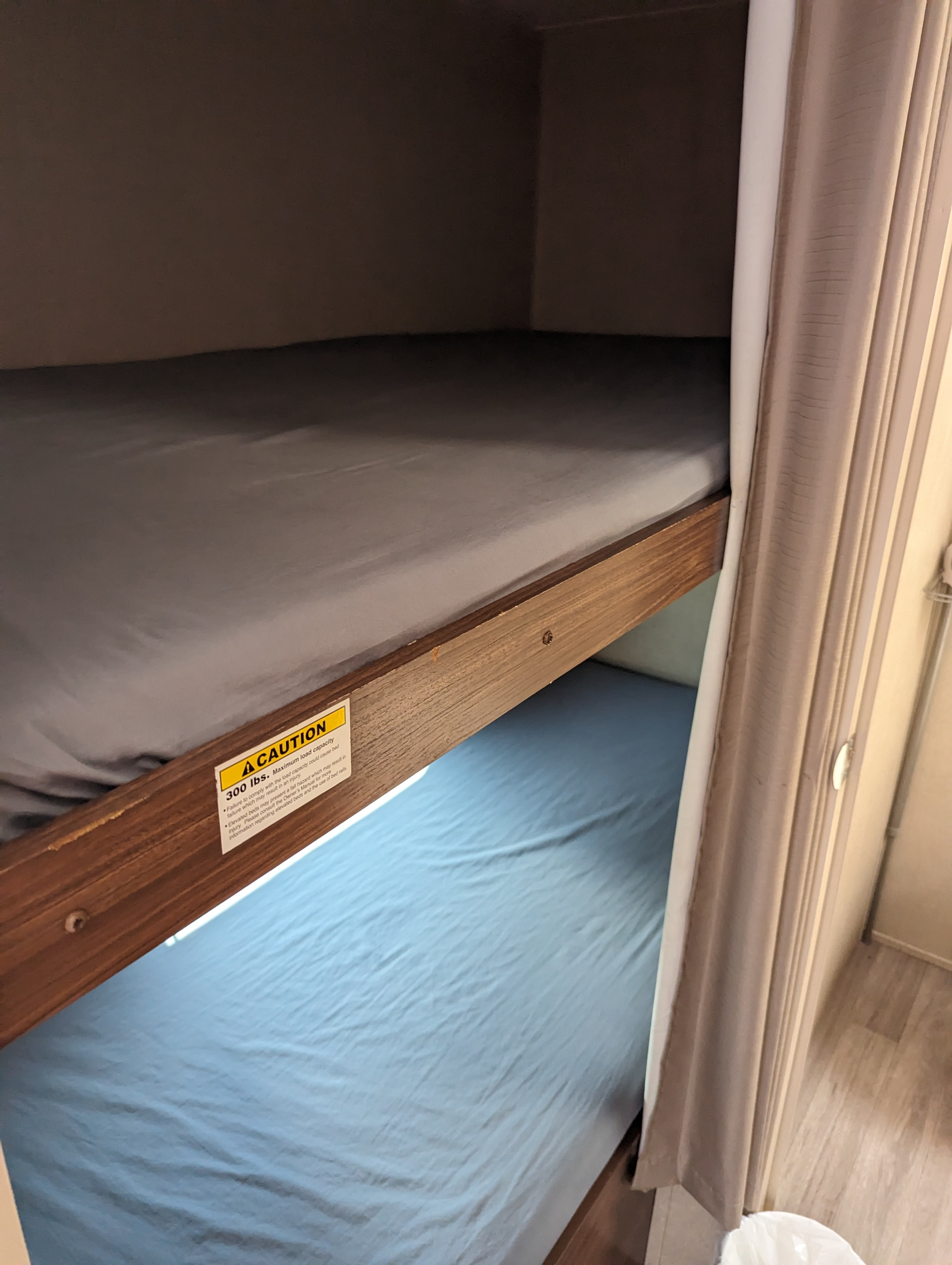 Inside an RV rental, bunk beds are made with gray and blue sheets. A caution sign on the top bunk warns of a 300 lbs weight limit.. Dutchmen Coleman Lantern LT 2017
